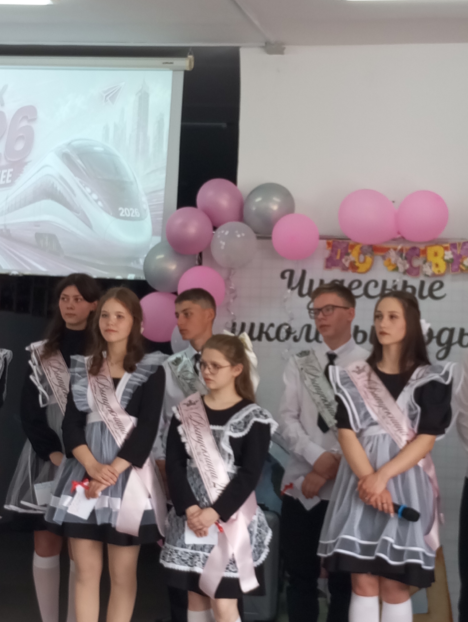 Captured in the Russian city of Буренка, the image presents a group of six young people standing on a stage. They are wearing traditional school uniforms and graduation sashes, suggesting a graduation ceremony. Balloons, both pink and gray, are arranged behind them. A banner displaying celebratory text is also visible. A screen in the background shows an image of a high-speed train, marked with "2026". The individuals are mostly looking forward, with neutral expressions. One of the girls is holding a microphone. They all seem to be waiting for something or listening.