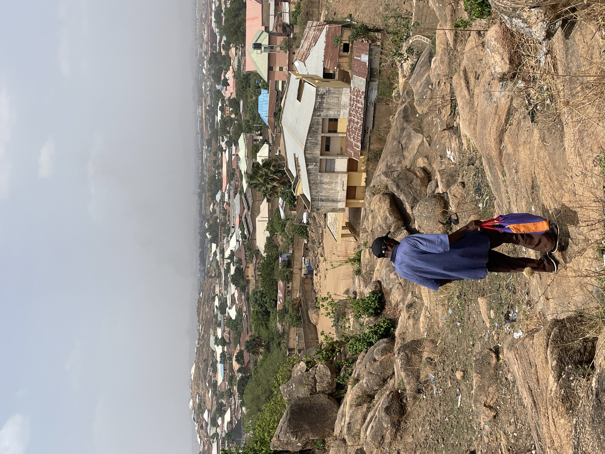 A slightly tilted, eye-level, outdoor shot in daylight depicts an African man on a rocky hillside overlooking a cityscape, which is located in the city of Jos, Nigeria. The man is wearing a navy-blue t-shirt, brown pants, black shoes with white soles, and a black baseball cap. He is holding a blue and orange bag. The hillside is covered with dry grass, rocks, and trash. In the background, there are many buildings. A hazy sky can be seen in the background.