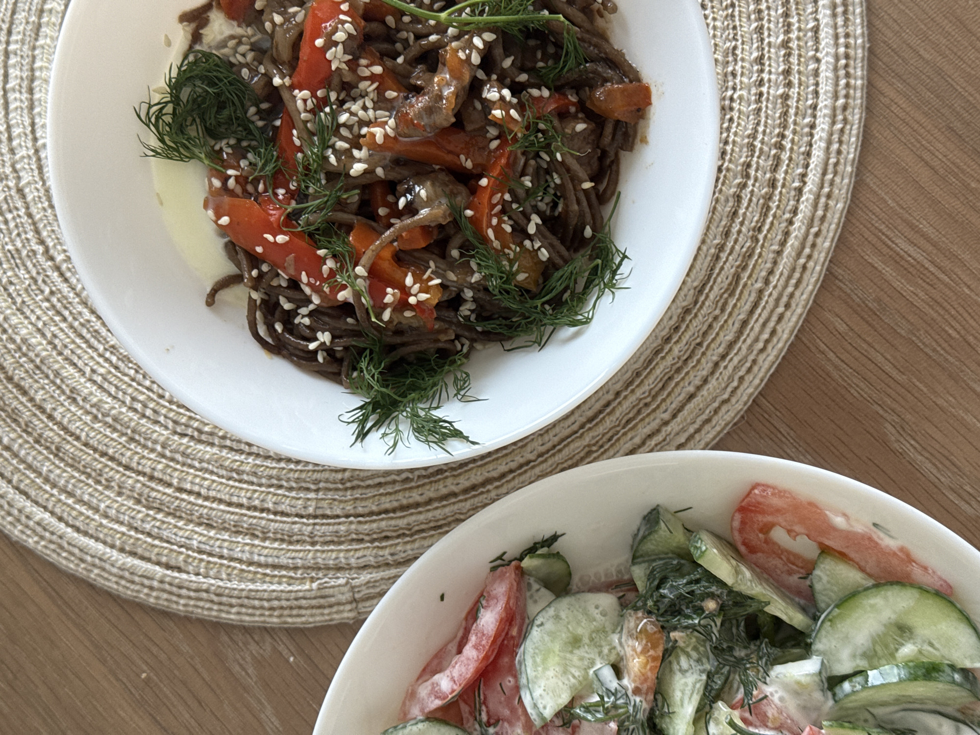 The image shows a still life of two white bowls of food on a light brown woven placemat that rests on a light brown wooden table. The bowl at the top contains dark noodles, red and brown peppers, seeds, and green sprigs of dill. The bowl at the bottom contains sliced cucumbers and tomatoes with a creamy dressing, garnished with dill. The location is likely a home dining setting, but the exact city or location in Shirkovo, Russia cannot be determined from the image.