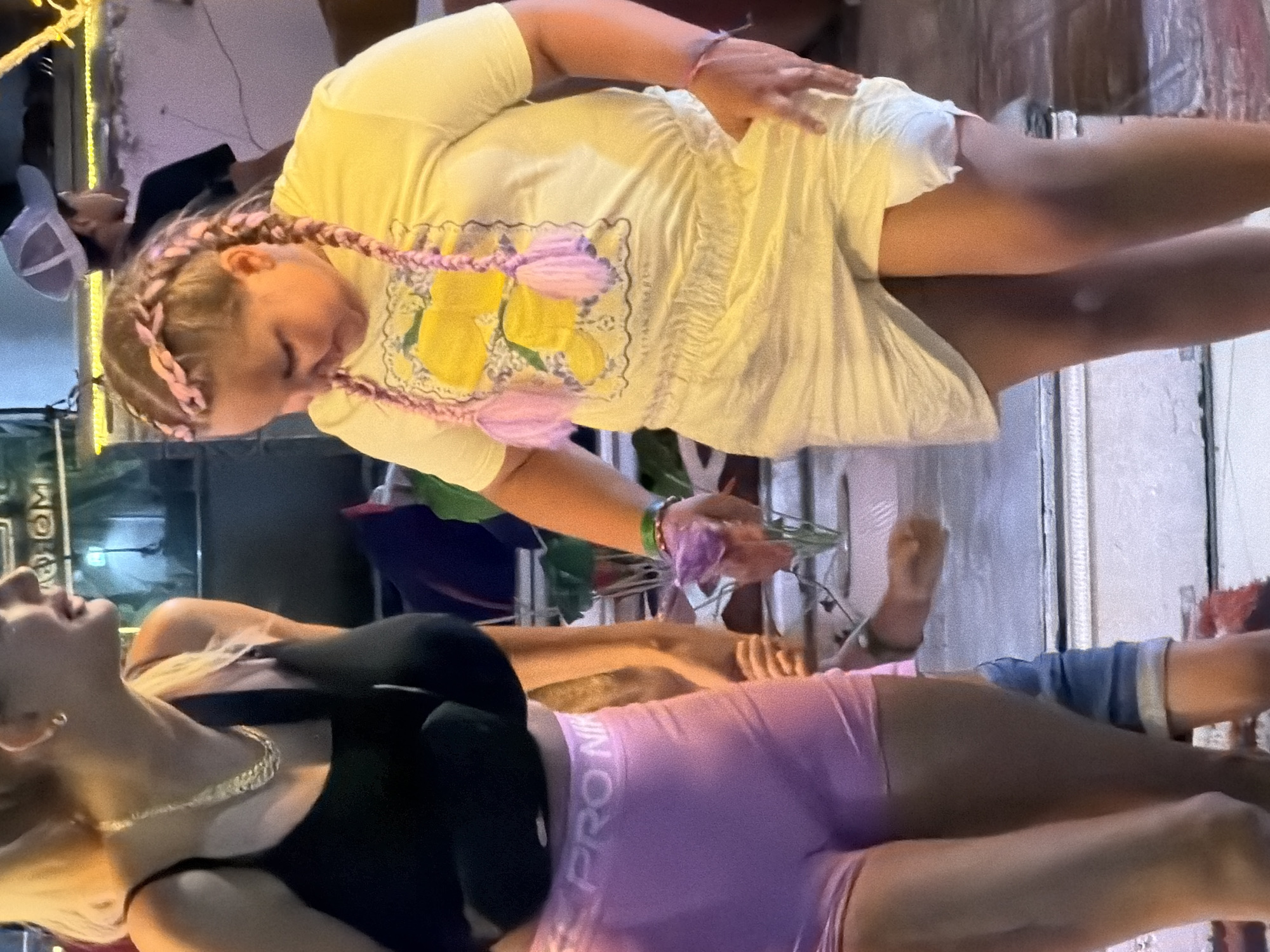 The image captures a scene in Sharm El Sheikh, Egypt, likely at a street market or outdoor dining area. In the foreground, a young girl with blonde hair styled in pink braids and adorned with pink tassels is wearing a yellow t-shirt with a lemon graphic and a white skirt. She is looking down and holding a purple object. To her left, a woman with blonde hair is wearing a black tank top and light pink Nike shorts. She is also holding a purple object and looking to her right. Partially visible in the background are other people, including a person wearing a white baseball cap and a dark t-shirt, and a child in denim shorts. The environment appears to be outdoors with some artificial lighting, suggesting an evening or nighttime setting. The ground surface is made of concrete or stone, with some steps or tiered platforms visible. Greenery and decorative elements are present, hinting at a tourist-oriented location.