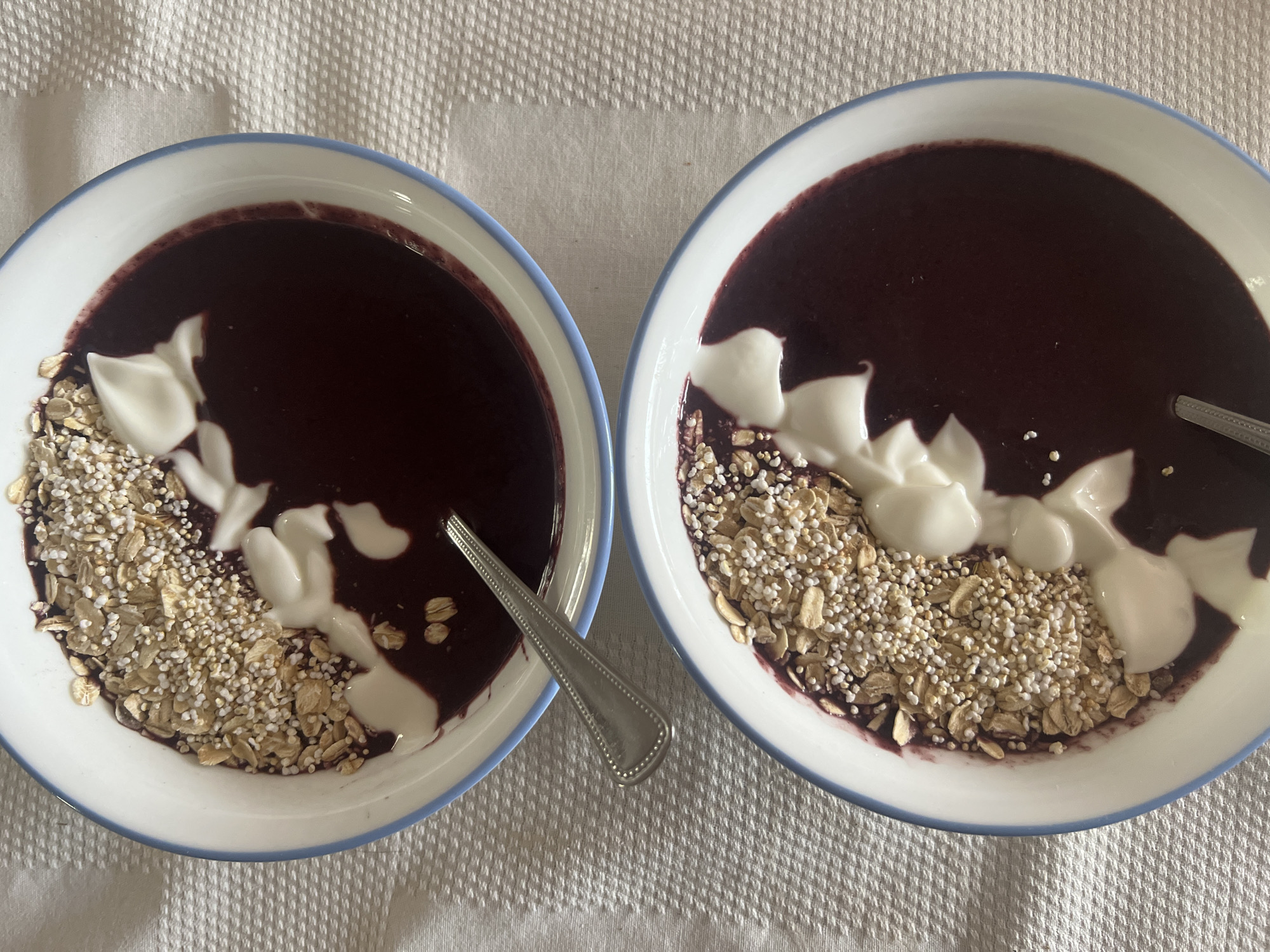 Two bowls of dark red smoothie are presented on a white, textured tablecloth. Each bowl, trimmed with light blue, contains a portion of what appears to be yogurt, and a mixture of oats and popped grains. A silver spoon rests in one of the bowls. The location is likely a home setting; there is no evidence to suggest it is Mexico City.