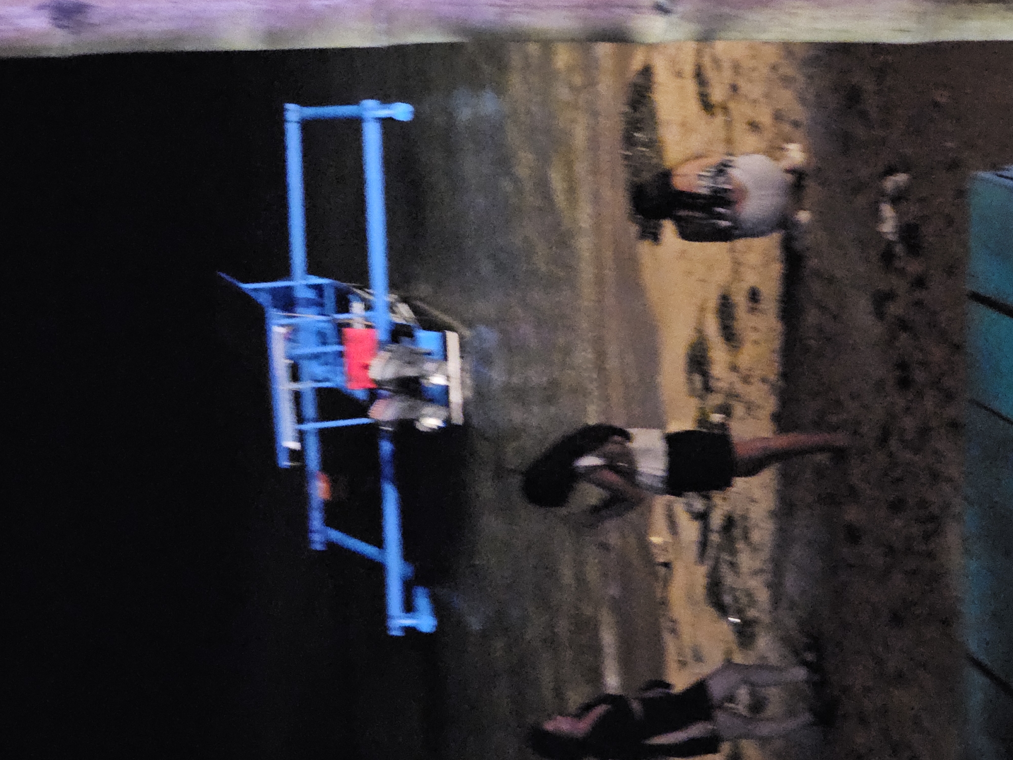 The image depicts a dimly lit scene at what appears to be a beach in Denpasar, Indonesia. In the background, a blue and red boat is partially submerged in dark water. In the foreground, three individuals are visible on the sandy shore. To the left, a person in dark clothing is seen from the side. In the center, another individual with dark hair is facing away from the camera, wearing a white top and a dark skirt. To the right, a third person is seated on the sand, facing away, wearing a patterned top and white bottoms. The overall environment is dark, suggesting it is nighttime.