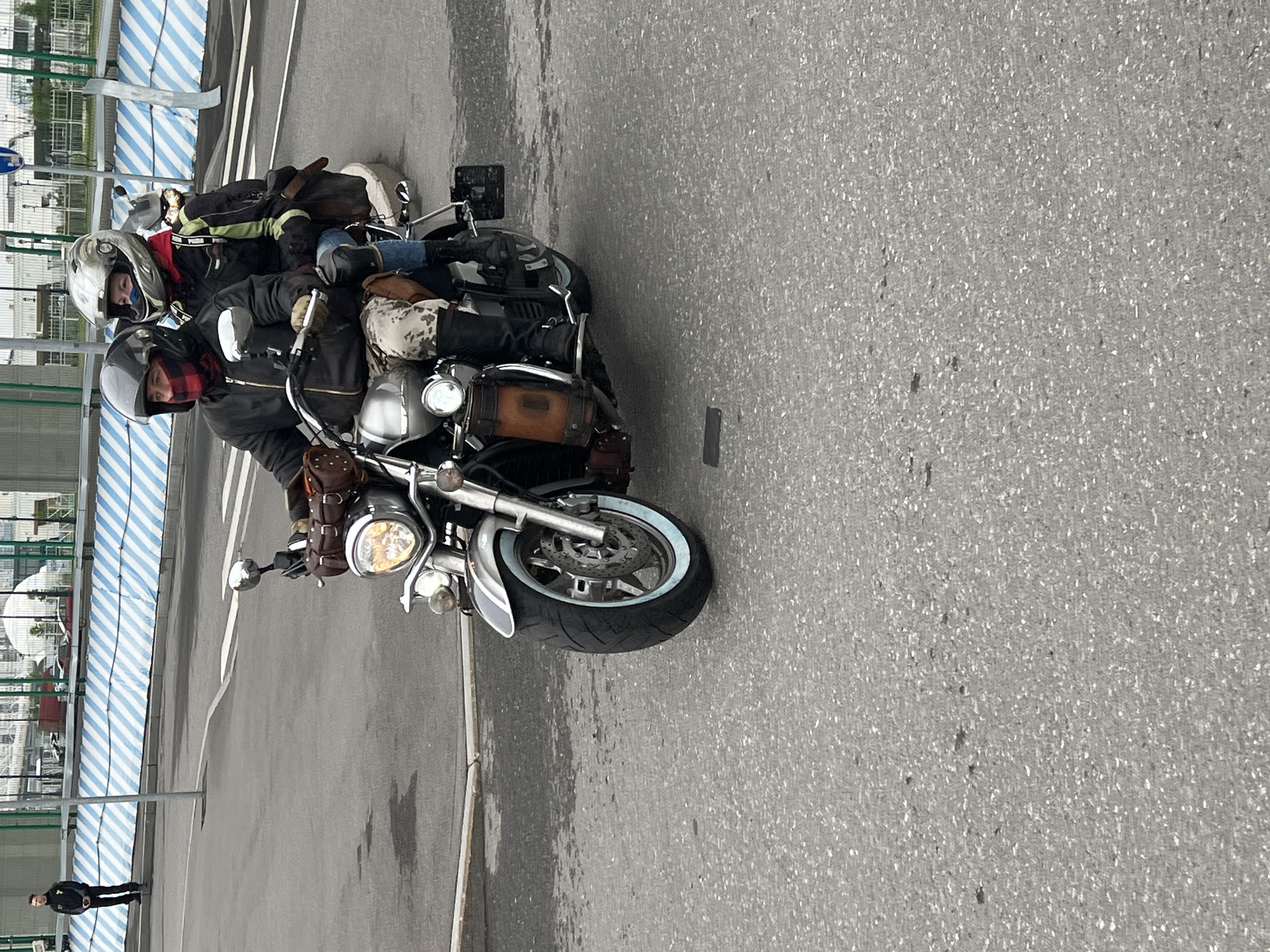 Two individuals are seated on a motorcycle. The motorcycle is parked on an asphalt surface. A person stands in the background near a blue and white striped structure. The location is Saint Petersburg, Russia.
