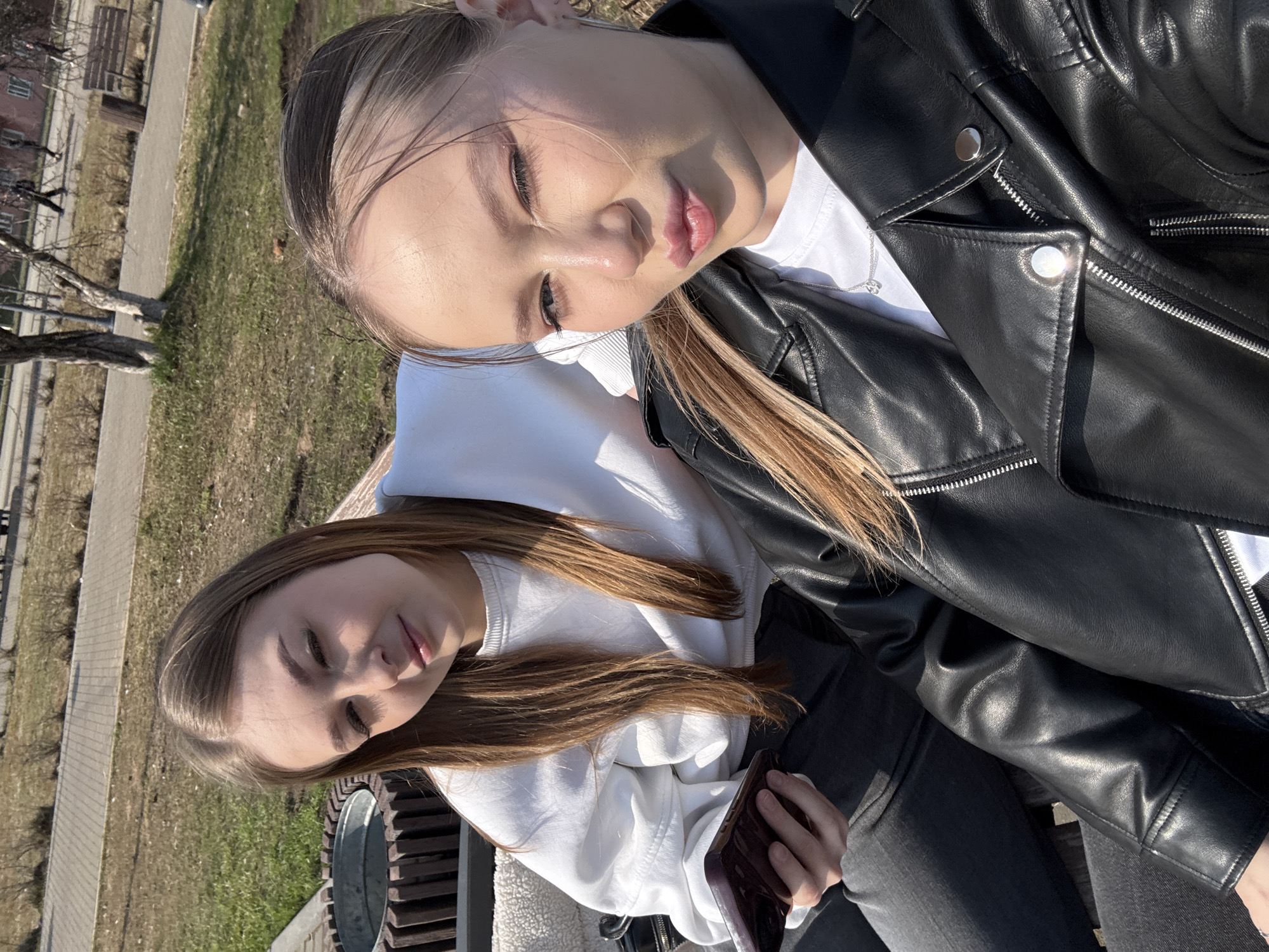 Two young women are sitting outdoors on a park bench. One woman, wearing a white sweatshirt, sits on the left side, holding a cell phone in her left hand. The other woman, wearing a black leather jacket over a white T-shirt, sits on the right side. They are close together, possibly leaning on each other.
In the background, there is a grassy area and a sidewalk. Further in the background, there is a building and trees, suggesting an urban or suburban setting. Based on the location information, the scene is in Chaikovsky, Russia.