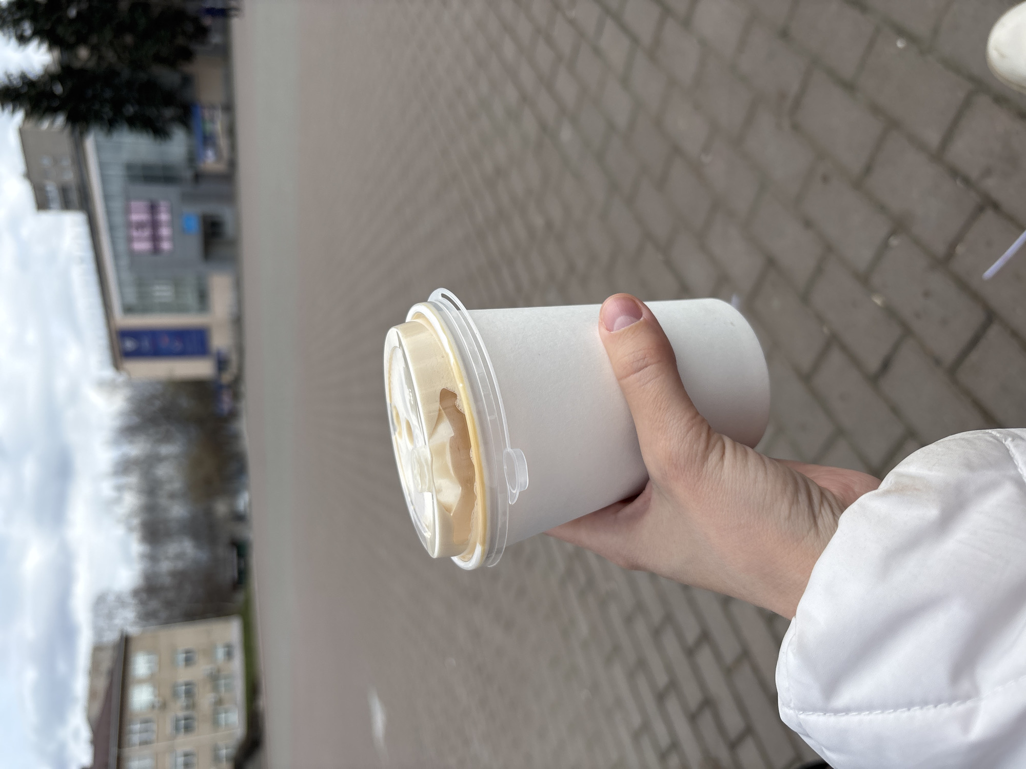 A hand holds a sealed paper cup containing a light-colored liquid, presumably a drink, on a paved street in Chaikovsky, Russia. In the background, a building with multiple windows and a few trees is visible. The sky is overcast. The sleeve of a white jacket is also visible in the lower right of the frame. The pattern of the street is of elongated bricks. The building in the background is blurred.
