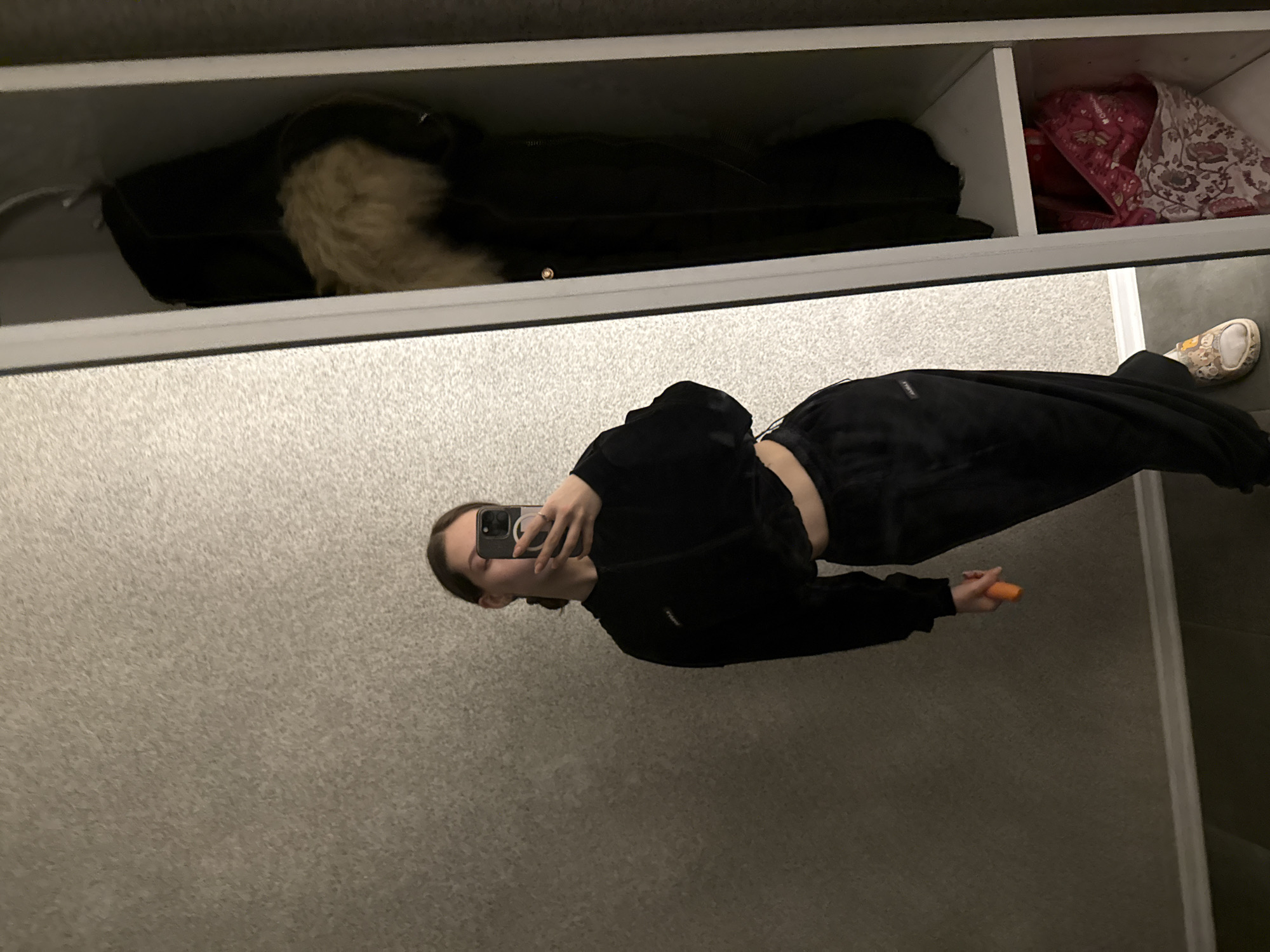 The image is a selfie taken in a mirrored closet. A woman in black sportswear with her midriff exposed holds a phone in her left hand and a small orange cylinder in her right. She is standing in front of a closet with items on the shelf: a black coat with fur trim and some folded fabrics with floral patterns. The background is a light gray textured surface. The exact location cannot be determined from the image, as there are no landmarks or other features indicating Sofia, Bulgaria.