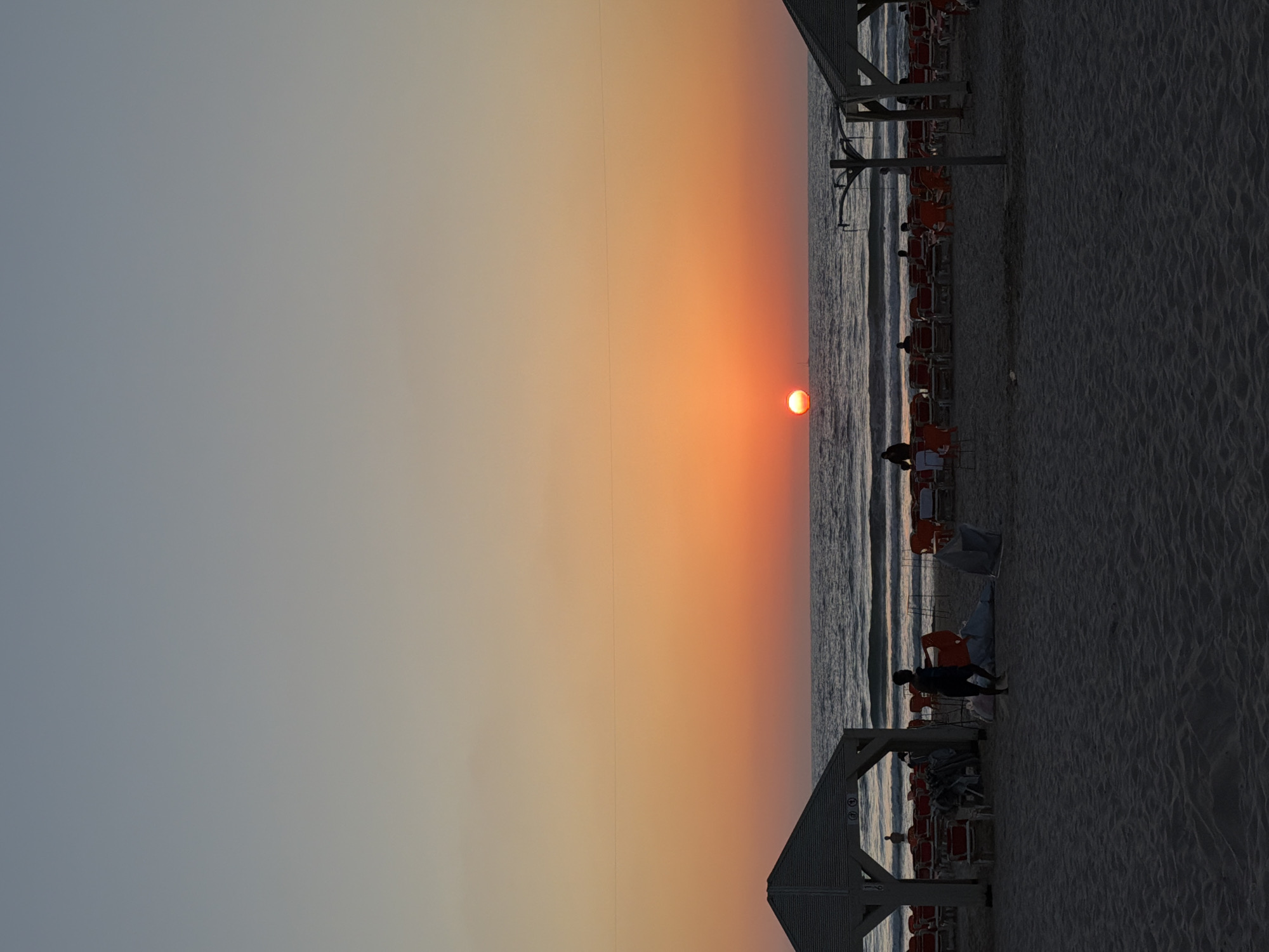 The image depicts a sunset at the beach in Tel Aviv, Israel. The sun is positioned on the horizon, creating a gradient of orange and yellow hues in the sky. The sea is visible between the sky and the beach, with gentle waves rolling in. There are people visible on the beach. There are umbrellas and chairs on the beach. The sand appears to be a fine texture. The image is slightly vertically oriented.