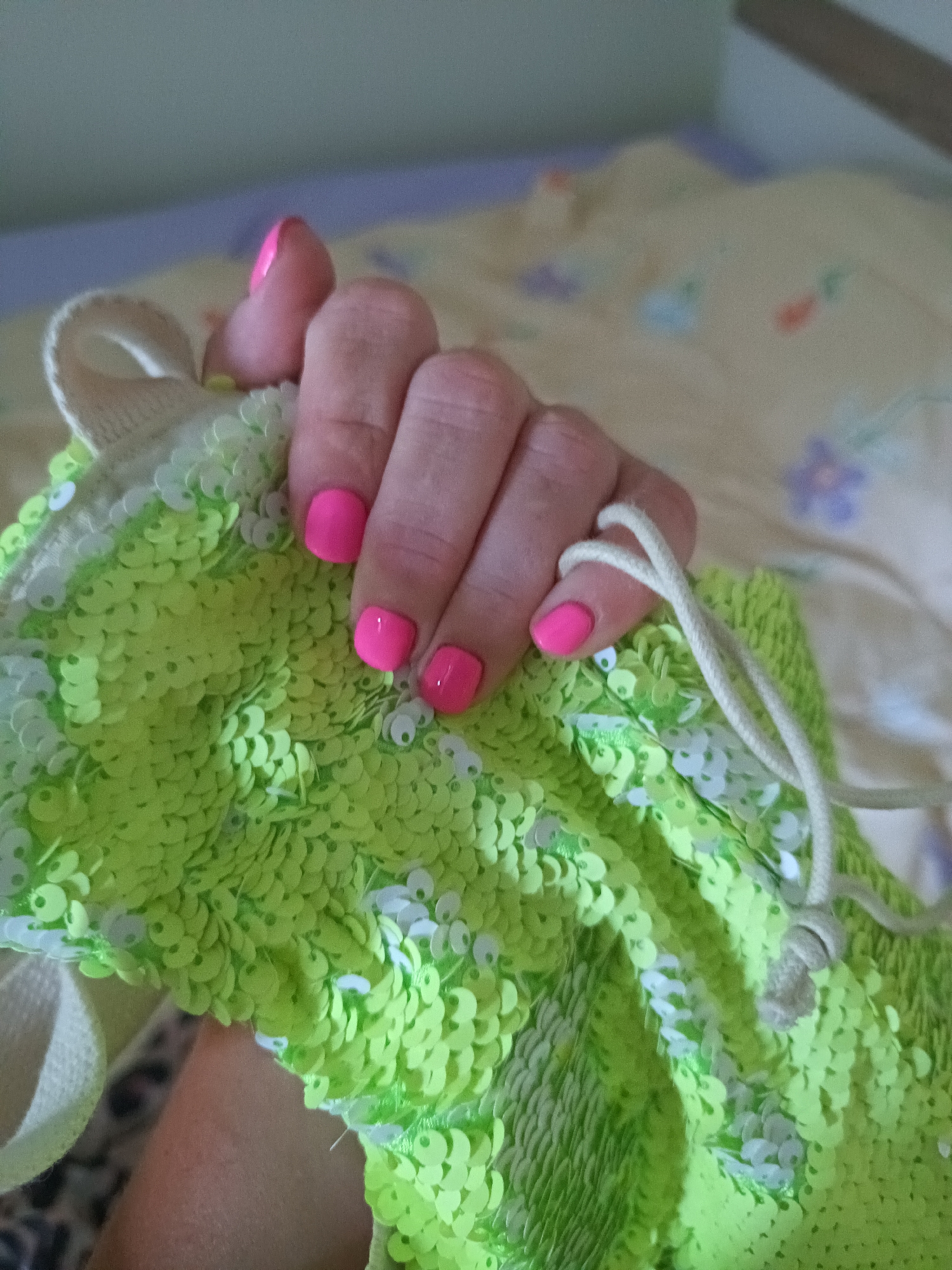 A hand with pink nail polish holds a green sequined drawstring bag against a blurred background.