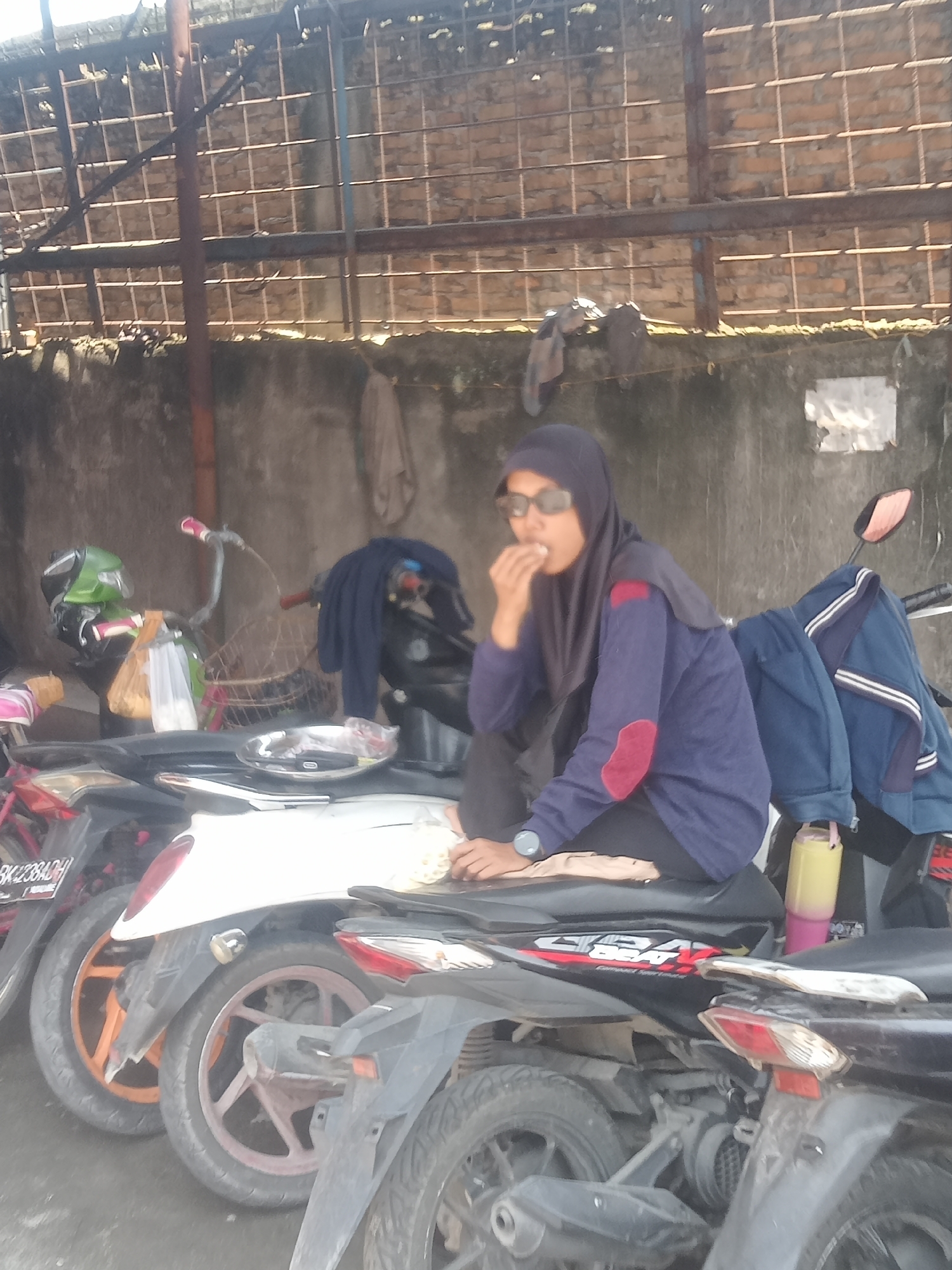 In Sunggal, Indonesia, a young woman wearing a dark hijab and sunglasses sits on a parked motorcycle, eating something from her hand. She is dressed in a long-sleeved navy blue shirt with a red patch on the left elbow. She is positioned next to other motorcycles, one of which is white and has an orange wheel rim. A blue jacket hangs on the handlebars of another motorcycle behind her. The scene takes place outdoors, possibly in a parking area, with a concrete wall in the background and a metal-barred roof overhead. The overall environment appears to be functional and somewhat utilitarian.