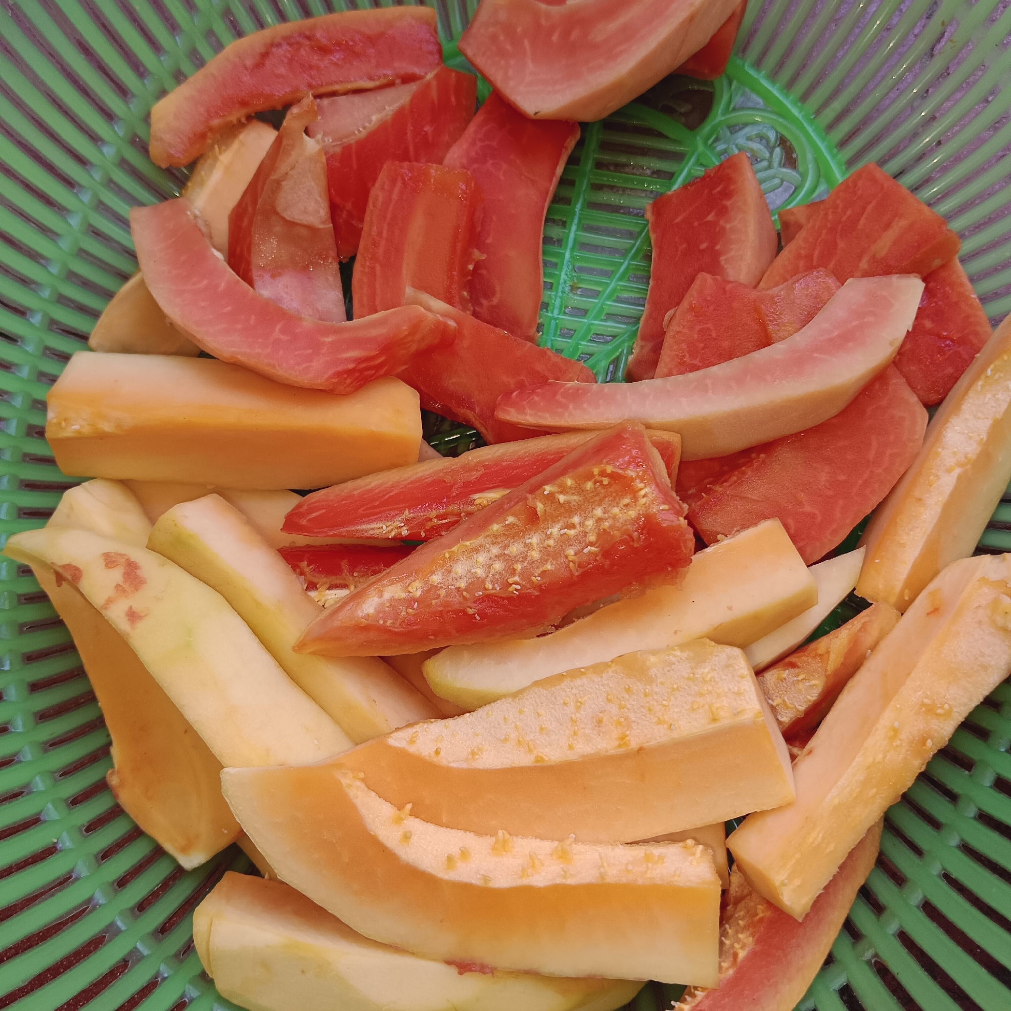 A close-up, top-down shot showcases a vibrant collection of sliced papaya fruit nestled within a green plastic basket. The fruit is cut into various shapes and sizes, ranging from wedges to long strips, with distinct variations in color, from deep reddish-orange to pale yellow-orange. Some pieces reveal the speckled texture of the papaya seeds, while others are smooth flesh. The green basket's crisscross pattern is visible beneath and around the fruit, suggesting an outdoor setting, possibly a market or a home kitchen in Paupanda, Indonesia, where fresh produce is prepared.