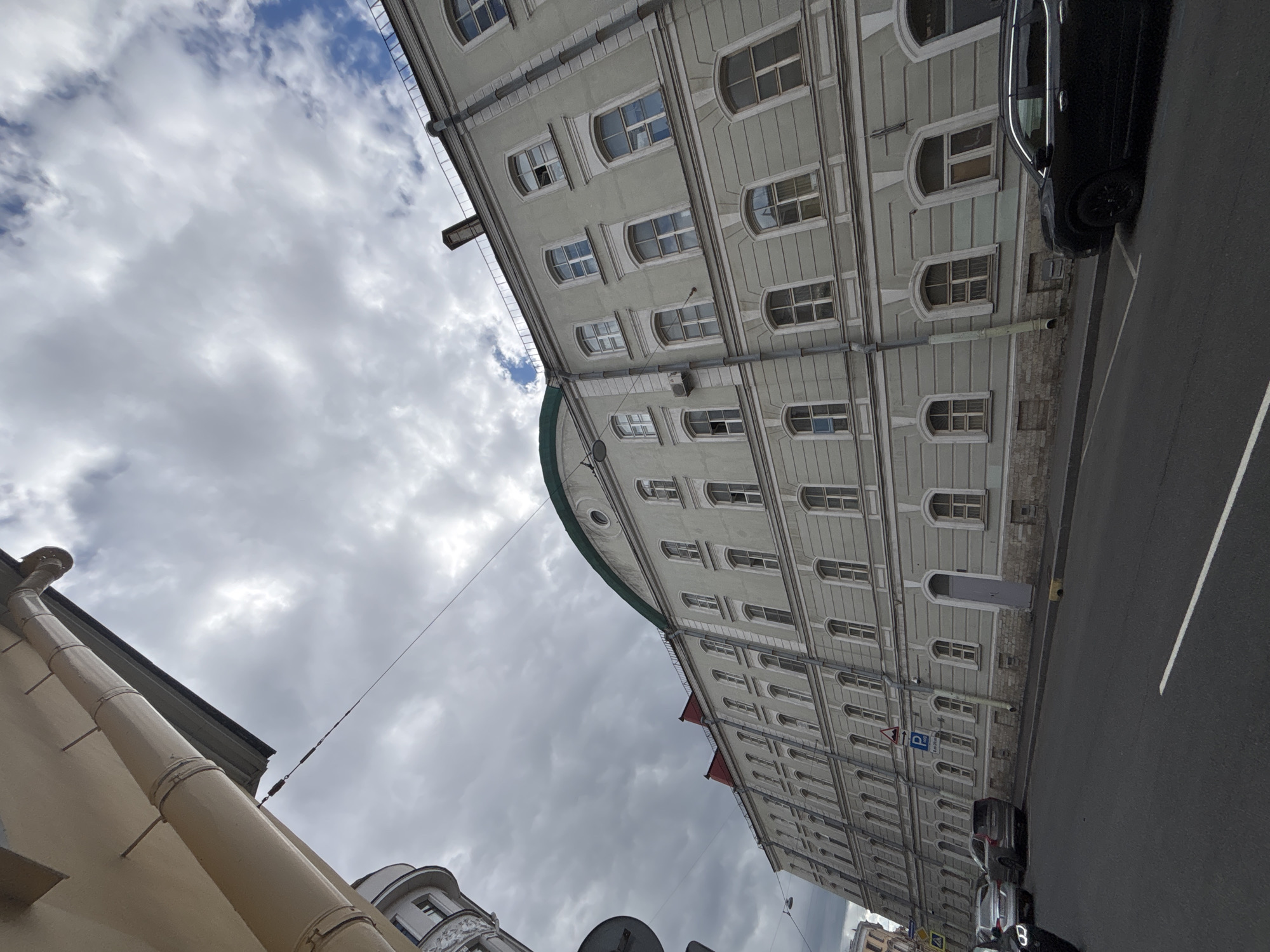 The image shows a wide, low-angle shot of a light-colored, multi-story building in Saint Petersburg, Russia. The building has a long facade with numerous windows, many of which are arched. The upper portion of the building features a green, curved roof. A downspout runs vertically along the side of the building. In the foreground, a street with a white lane marker is visible, with parked cars on the right side of the road. A black SUV is prominently parked on the right. The sky above is filled with dramatic, cloudy formations, with patches of blue sky visible. Another, older-looking building with columns is partially visible on the left edge of the frame, and a traffic sign with a blue 'P' and arrows is mounted on the facade of the main building. The overall impression is of an urban environment with a mix of architectural styles under an overcast sky.