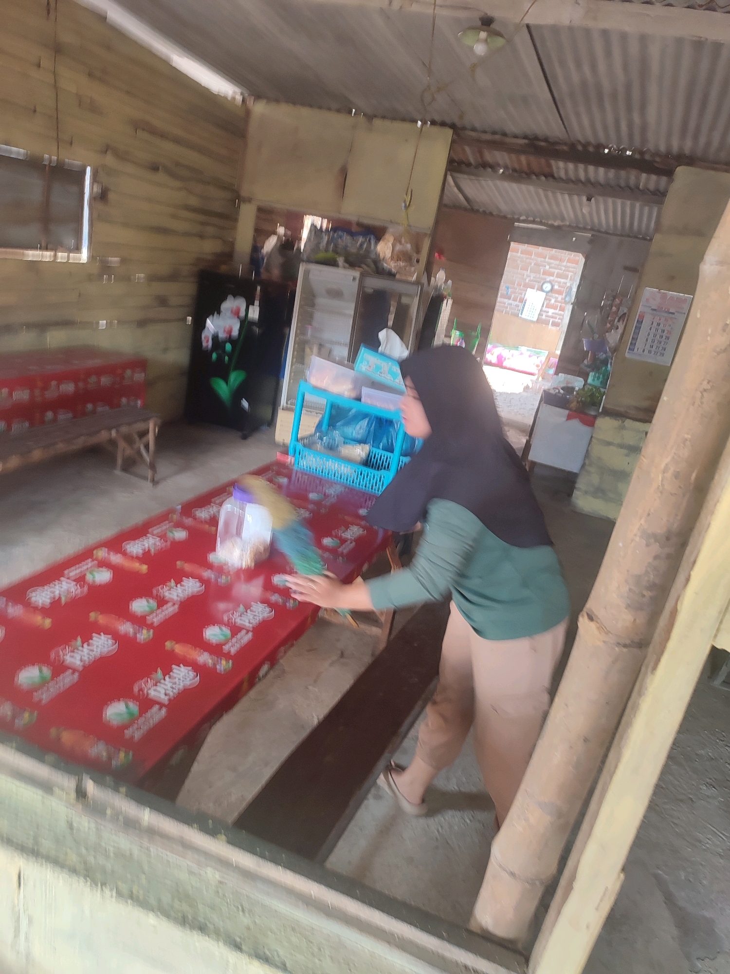 The image depicts an indoor scene, likely a simple dwelling or shop in Ringinanom, Indonesia. A woman wearing a black hijab and a green shirt is standing and cleaning a red table covered with a patterned tablecloth. She is holding a feather duster in her right hand and appears to be in the process of wiping the table. The table is set with a plastic bottle containing some sort of granular substance, possibly food items or ingredients.
The environment suggests a rustic setting. The walls are constructed from wooden planks, and the ceiling appears to be corrugated metal. In the background, various items are visible, including a black refrigerator with floral motifs, blue plastic shelves filled with containers, and a calendar on the wall. Sunlight filters in from an unseen source, illuminating the scene and casting some shadows. To the right, a large wooden beam or pillar is prominent, possibly part of the building's structure or a decorative element. The floor is concrete. The overall impression is one of a functional, unpretentious space where daily chores are being performed.