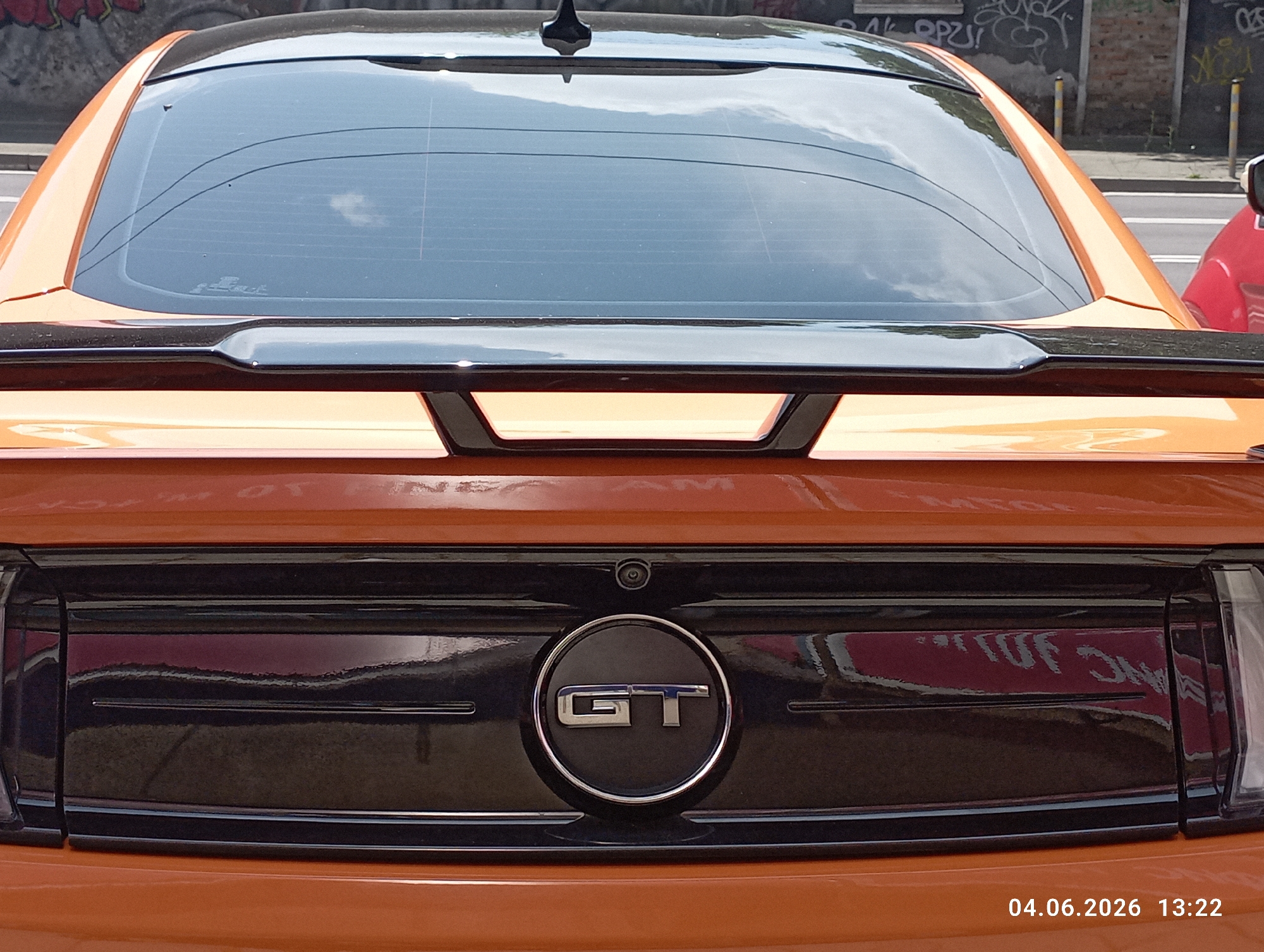 This media shows the rear view of a sporty, orange-colored car, identifiable as a Ford Mustang GT by the circular "GT" emblem centered on its trunk lid. The car features a black spoiler and a dark-tinted rear window reflecting the sky with clouds and overhead wires. A small black antenna is visible on the roof towards the top of the image.
Above the "GT" emblem, a small black backup camera is integrated into the trunk design. The lower part of the trunk and bumper area are black, reflecting indistinct shapes, including parts of a building, some red object (possibly another car), and hints of graffiti.
In the background, a brick wall covered with various graffiti is visible, along with yellow bollards and a suggestion of a road or sidewalk. The car appears to be stationary in an urban environment. The lighting suggests it is daytime with a clear or partly cloudy sky.
A digital timestamp "04.06.2026 13:22" is visible in the bottom right corner of the original image.