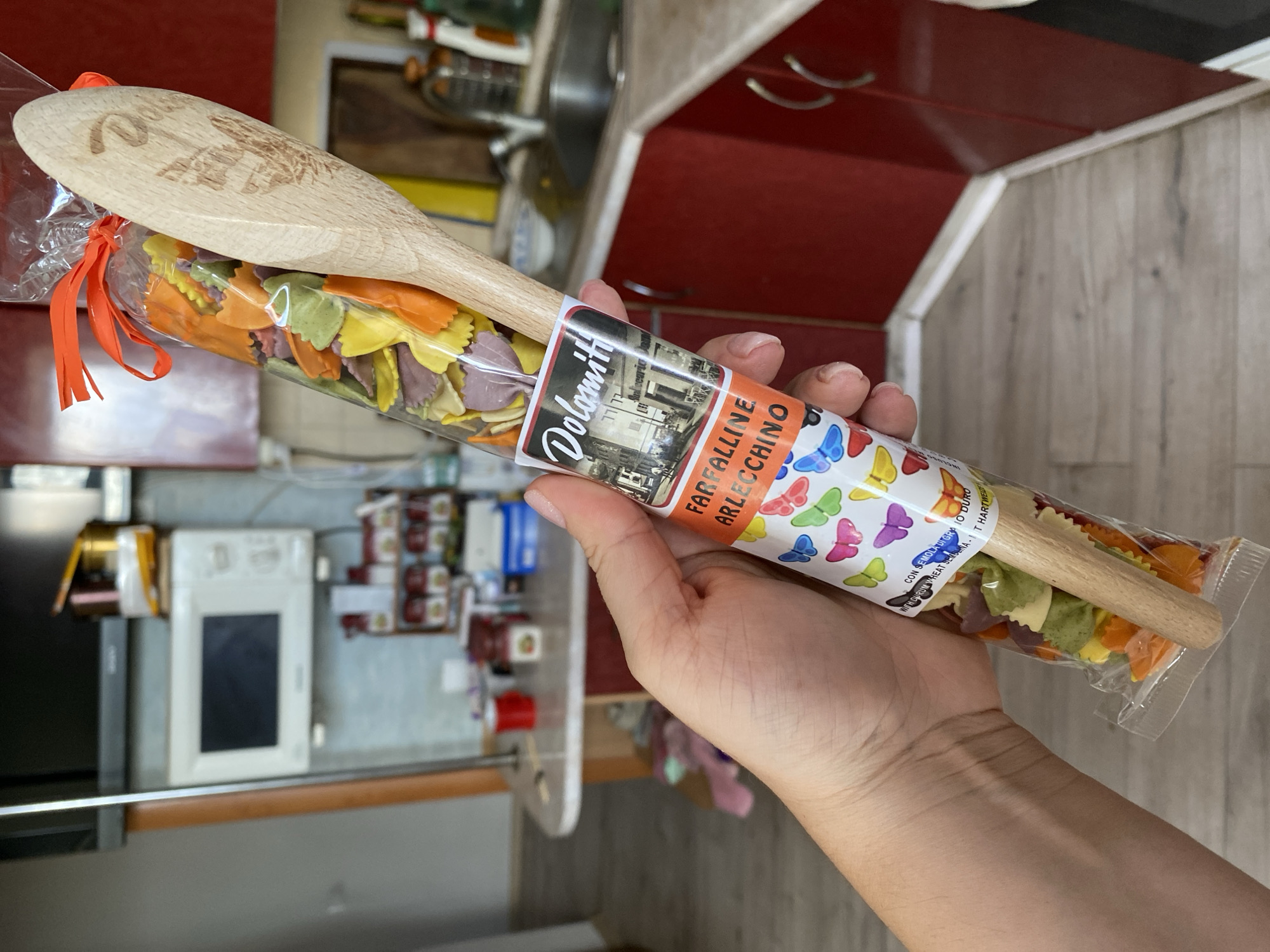 A hand is holding a clear plastic package of colorful farfalle pasta, each piece shaped like a bow tie, in assorted pastel colors of green, yellow, purple, and orange. A wooden spoon is inserted vertically into the package. A label on the package reads "Dolomiti," with an image of a building and text that appears to be "Farfalline Arlecchino." Below this, a pattern of colorful butterflies adorns the label. The background shows a kitchen environment with red cabinets, a microwave mounted on the wall, and a countertop with various items. The flooring is light-colored wood laminate. The image is taken in Molodizhne, Ukraine.