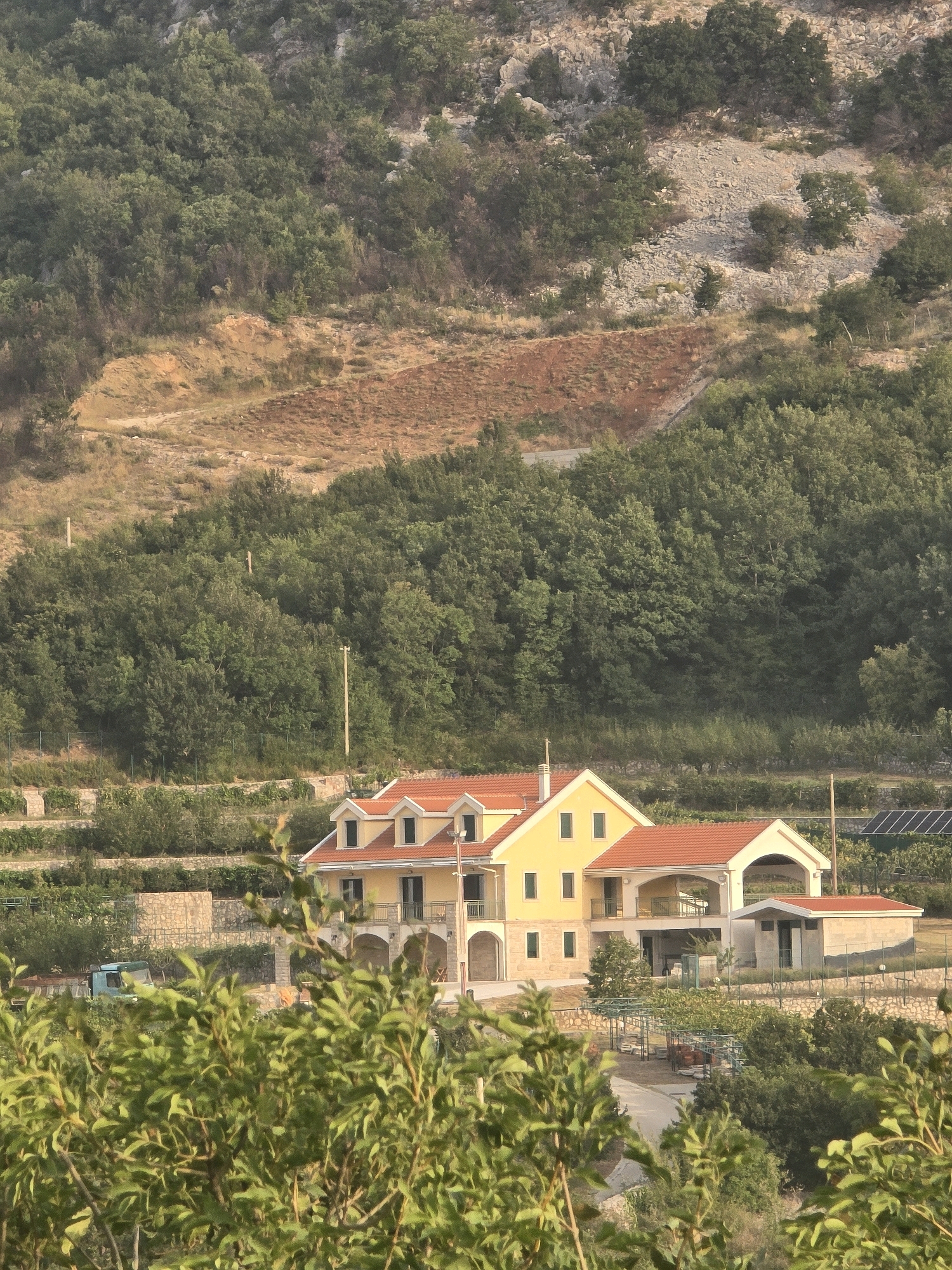 In the city of Gornja Vrbica, Montenegro, a large, multi-story villa with a red-tiled roof is situated on a hillside. The villa is painted a pale yellow and features several balconies and arched doorways. Adjacent to the villa is a smaller, single-story structure with a similar red-tiled roof. The property is surrounded by lush green trees and vegetation, with terraced gardens visible in the background. A blue truck is parked near the villa. The terrain suggests a rural or semi-rural setting, with a rocky, wooded hillside rising behind the structures. Solar panels are visible on the grounds. The scene appears to be captured during daylight hours, with sunlight illuminating the buildings and landscape.