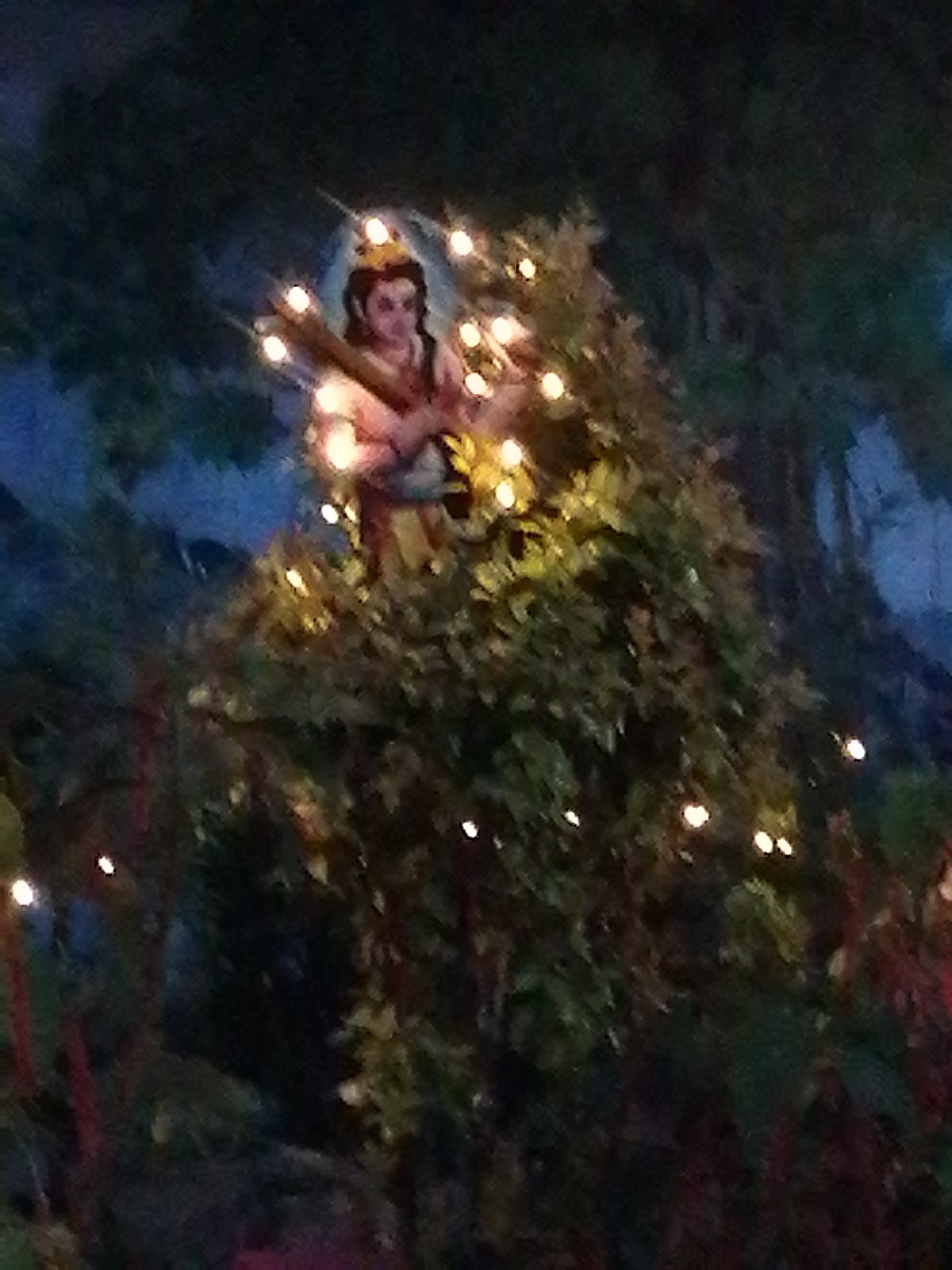 A person, appearing to be a religious figure, is the focal point of the image. This figure is positioned amidst dense foliage and surrounded by a string of small, warm-toned lights. The figure is holding a bundle of sticks or a wooden structure across their shoulders. The backdrop suggests an outdoor or forested environment, possibly a display within a larger setting. Considering the information about Da Lat, Vietnam, it is possible that this is a display within a religious site or a tourist attraction in that city. The scene has a slightly mystical or fantastical quality due to the combination of the religious figure, dense foliage, and the string lights.