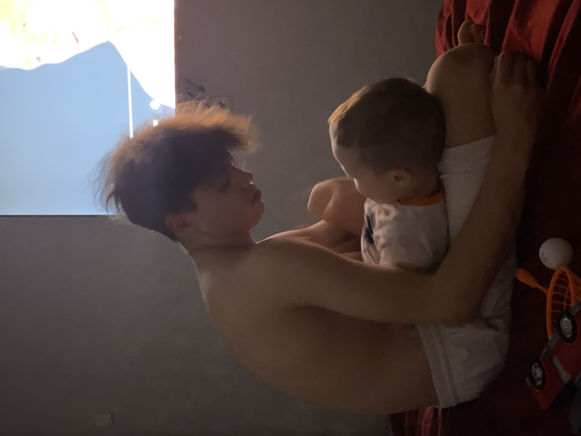 A young boy is holding a baby in his arms, sitting on a red surface that looks like a bed or couch. The older boy is shirtless, wearing white shorts. The baby is wearing a white shirt with an orange trim. To the right of the baby, there is a white ball, a toy tennis racket, and a toy car. There is a light colored wall behind the children, with a screen displaying a light blue image in the upper left corner. The location is not apparent from the image. The city of Sochi, Russia, is not identifiable from the image.