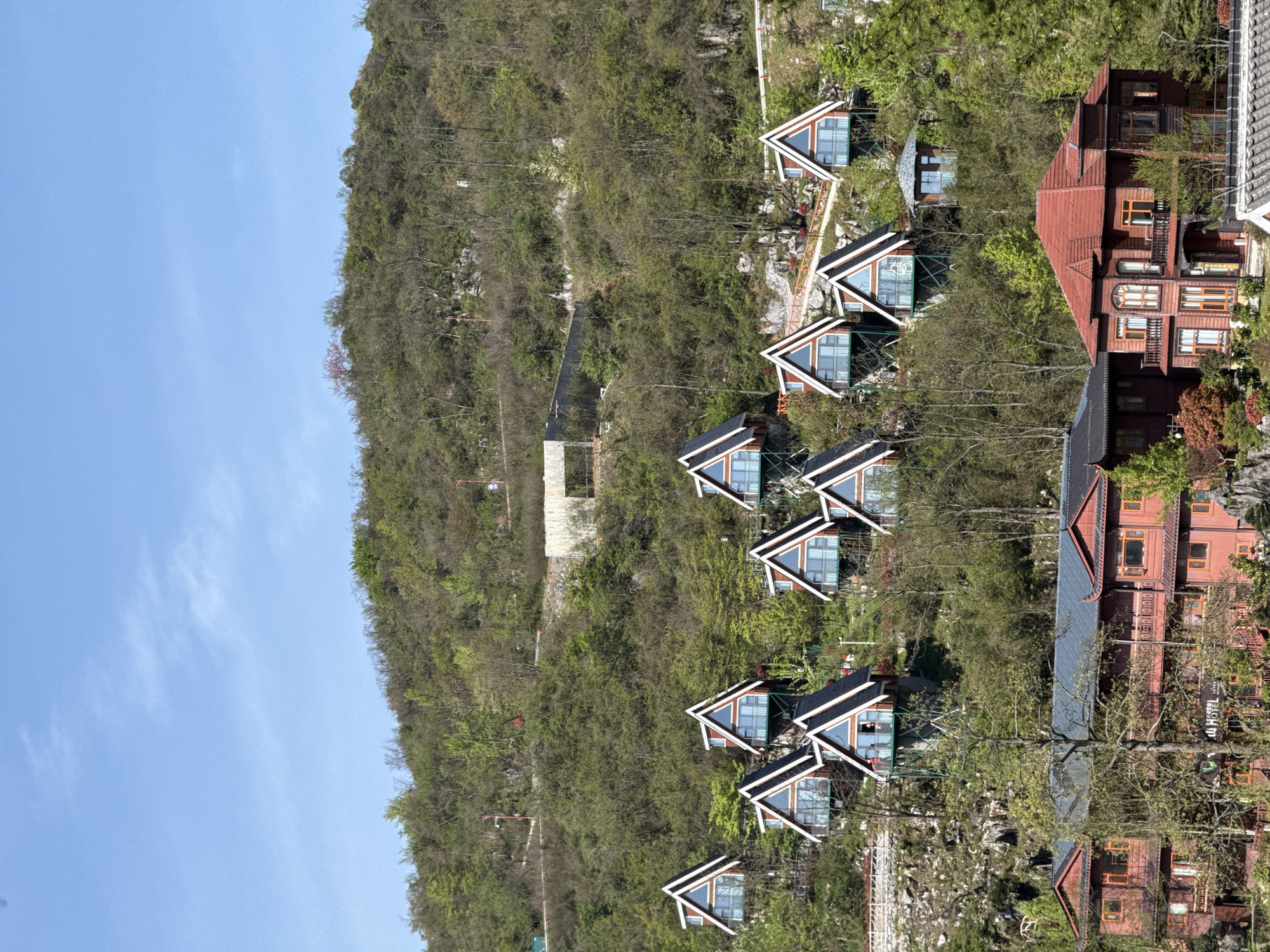 The image displays a picturesque hillside landscape, likely a resort or lodging area, under a bright blue sky with minimal clouds. The scene is dominated by a steep, tree-covered slope descending from left to right.
On the right side of the image, numerous buildings are strategically integrated into the verdant hillside. A series of modern A-frame cabins with large triangular glass fronts and dark, contrasting roofs with white trim are prominent. These structures appear to be individual units, possibly guest accommodations, and are connected by what seem to be pathways or architectural elements.
Further down and to the right, larger, multi-story buildings with a reddish-brown, possibly wooden, exterior and distinct red tiled roofs are visible. These larger structures feature multiple windows, some rectangular and others with arched designs, and appear to have balconies or outdoor access points. On one of these larger buildings, the text "LU HOTEL" is clearly visible, indicating its function.
The hillside itself is densely forested with a mix of mature trees and undergrowth, suggesting a rich natural environment. Some rocky outcrops are also visible among the vegetation. The time of day is clearly daytime, characterized by bright, even lighting and a clear sky, implying sunny weather. There is no discernible human activity captured in the frame. The overall impression is one of a serene retreat nestled within nature, consistent with a resort setting in Yongding District, China.
