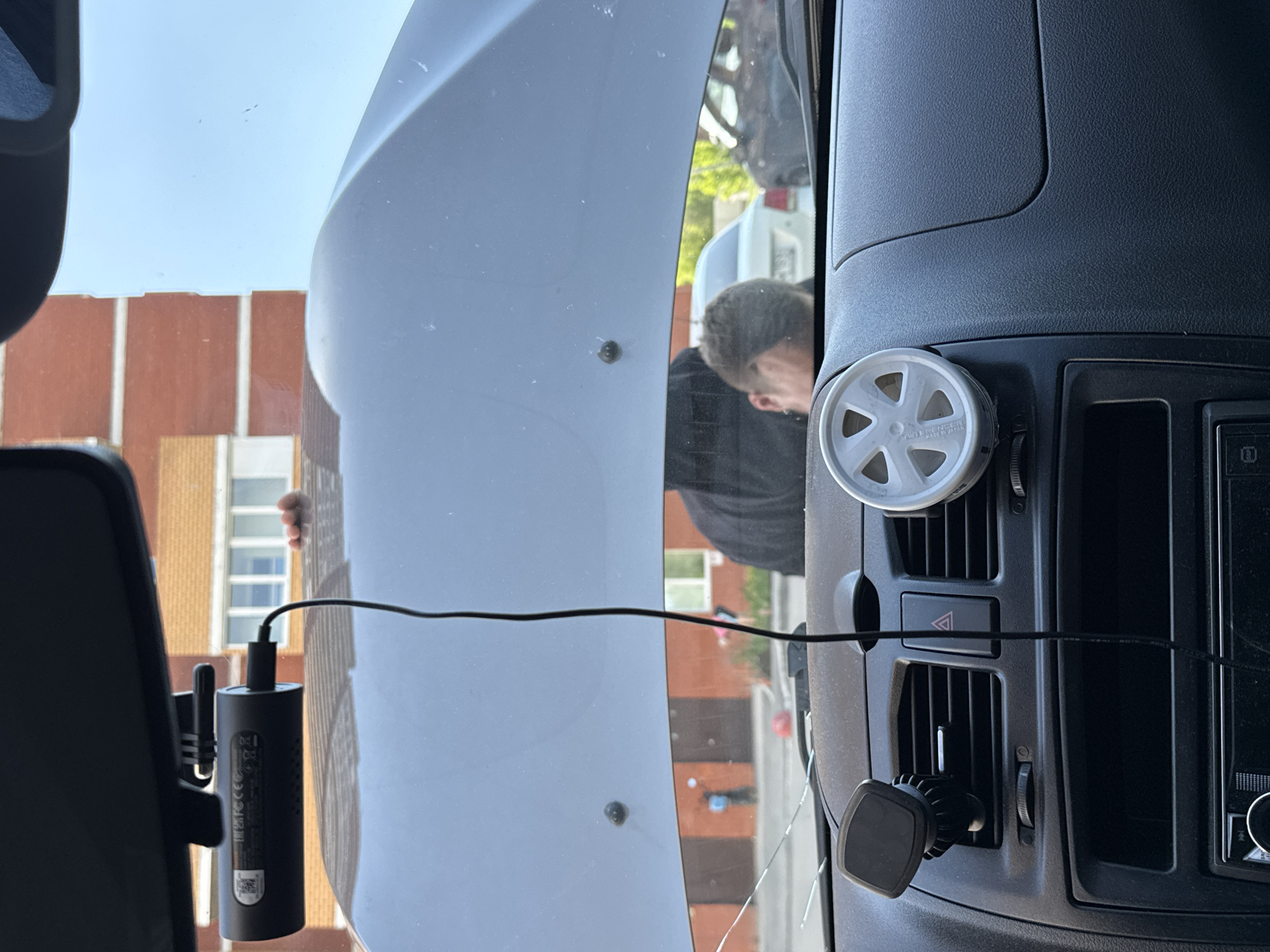 The view is from inside a vehicle, possibly a truck, looking out the windshield. A person is visible outside the vehicle, leaning toward the windshield. A building with red brick and white-framed windows is visible in the background, along with a blue sky. Inside the vehicle, a black electronic device is attached to the windshield with a wire running across the glass. The dashboard of the vehicle is visible, featuring air vents, a hazard light button, and a circular air freshener. A magnetic phone mount is attached to one of the air vents. The location is not discernible as Perm, Russia, based on the visual evidence.