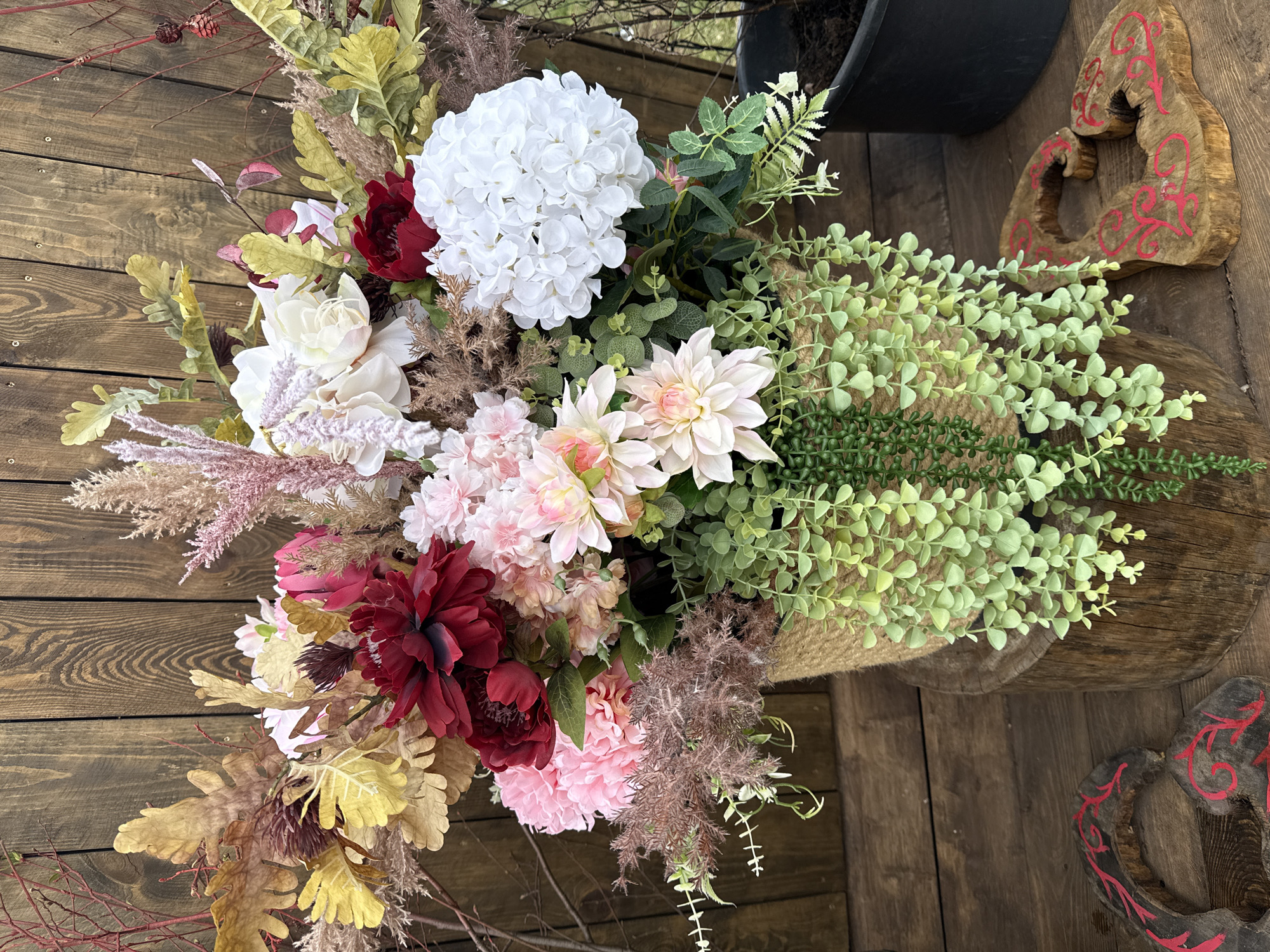 An overhead shot captures a vibrant, multi-textured floral arrangement set on a rustic wooden plank surface. The arrangement is a rich tapestry of artificial flowers and natural elements, primarily in shades of white, pink, red, green, and muted yellows and browns, suggesting an autumnal theme.
Dominant flowers include large clusters of white hydrangeas, white peonies or roses, light pink dahlias, deep burgundy peonies or dahlias, and various smaller pink blossoms. Complementing the flowers is an abundance of diverse foliage: bright green trailing plants, darker green fern-like leaves, muted yellow and brown oak-shaped leaves, and feathery pinkish-brown pampas grass. Bare reddish branches with small dried berries or seed pods also emerge from the arrangement.
The flowers are nestled within a rustic, possibly rope-wrapped or woven container, partially visible on the right side. The wooden surface beneath is a deck or floor with distinct grain patterns. In the upper right corner, a dark planter or pot is partially visible, along with a carved wooden decorative object shaped like an interlocking heart, adorned with red swirling patterns.
The scene is static, showcasing the intricate details of the floral display under what appears to be bright daylight in an outdoor setting. No specific activity is occurring, and there is no visible text. The overall impression is one of natural beauty and decorative artistry. This image is from Tyulyuk, Russia.