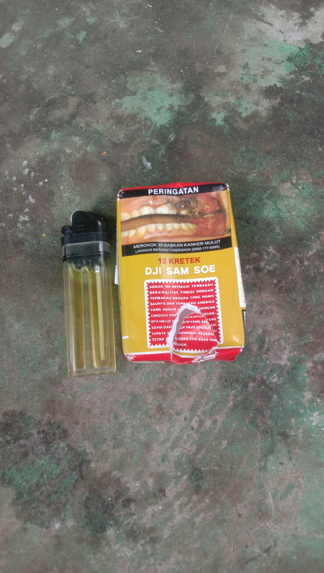 A transparent yellow plastic lighter with a black cap is positioned to the left of a yellow cigarette pack on a mottled grey and green concrete surface. The cigarette pack has a red border at the top, with the word "PERINGATAN" (Warning) printed in white. Below this, there is an image of a person's mouth, showing decaying teeth and a diseased gum. The pack also displays text in Indonesian, including "MEROKOK SEBABKAN KANKER MULUT" (Smoking causes mouth cancer) and a helpline number. Further down, it reads "12 KRETEK DJI SAM SOE" and lists some ingredients and qualities of the cigarettes. The cigarette pack appears to be torn along its bottom edge. The overall scene suggests an informal setting, possibly outdoors, given the nature of the concrete ground. The location in Karang Anyar, Indonesia, implies a specific regional context for the products and the environment.