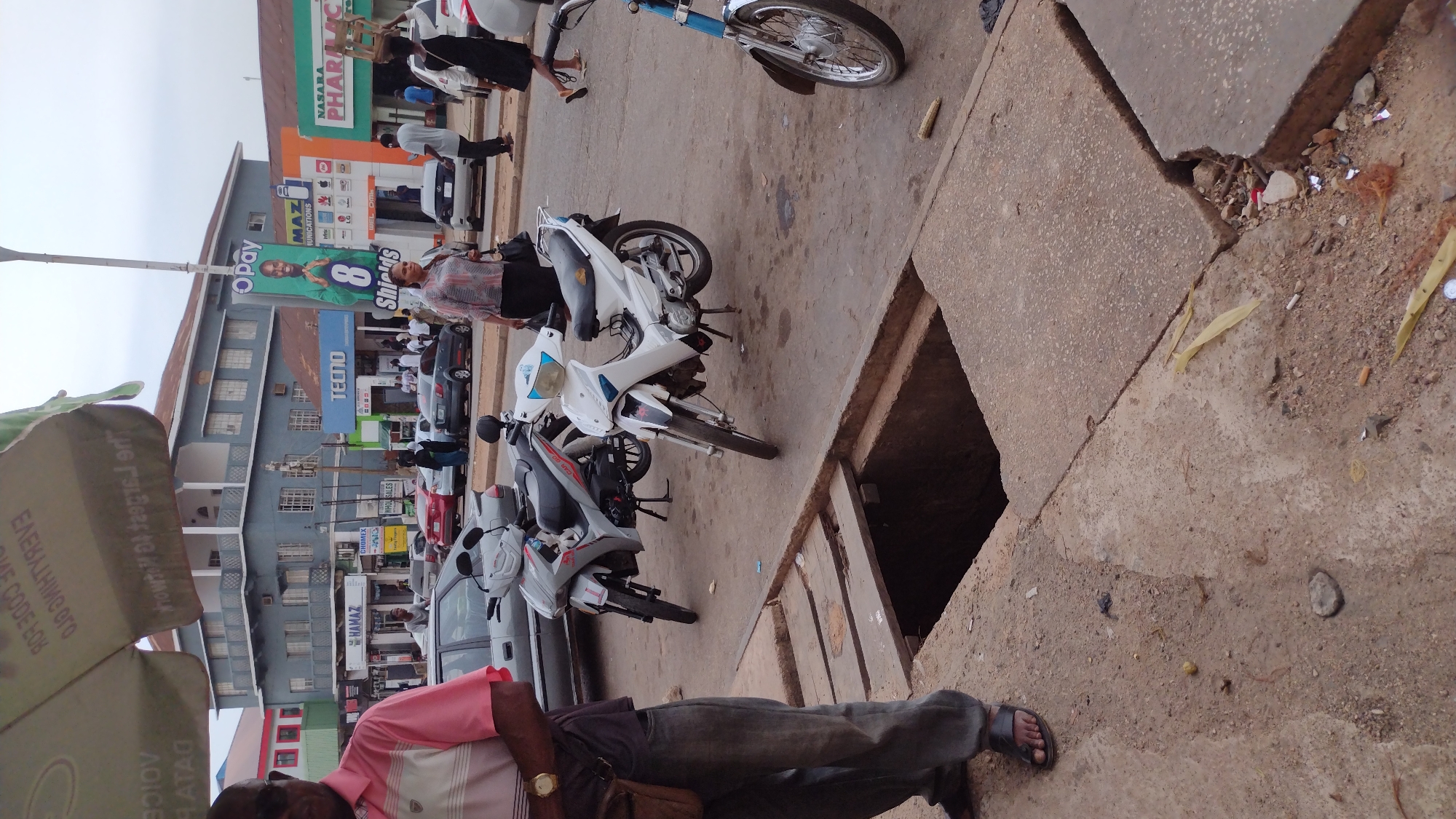 The image depicts a street scene in Jos, Nigeria. The foreground features a concrete sidewalk with an open, square drain. A man in a pink shirt and jeans stands near the drain, partially obscuring it. Several parked motorcycles line the street, their front tires pointing away from the camera. The building in the background has various commercial signs, including "Opay," "Tecno," "Hamaz," "Chomex," "Nasara Pharmacy," and "Shields." People can be seen walking along the sidewalk and in the street, including a woman walking past the motorcycles. The sky is overcast.