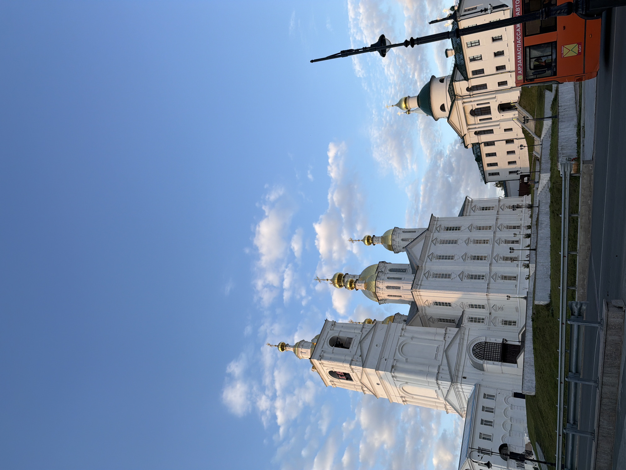 This is a full shot, taken outdoors, of a bright, white building with golden domes in Arzamas, Russia. The building appears to be a church or monastery, with a tall bell tower on the left and several smaller domes on the right. The architecture is ornate, with arched windows and decorative moldings. The building is situated on a slight incline, with a well-maintained grassy area in front of it. A road and a guardrail are visible in the foreground. To the right of the building, an orange bus is parked, with Russian text visible on its side. The sky is a clear blue with scattered white clouds, suggesting it is daytime. The lighting indicates it may be late afternoon or early morning, as there are some long shadows. No people are clearly visible in the image, though there might be a distant figure on the steps of the building on the right.