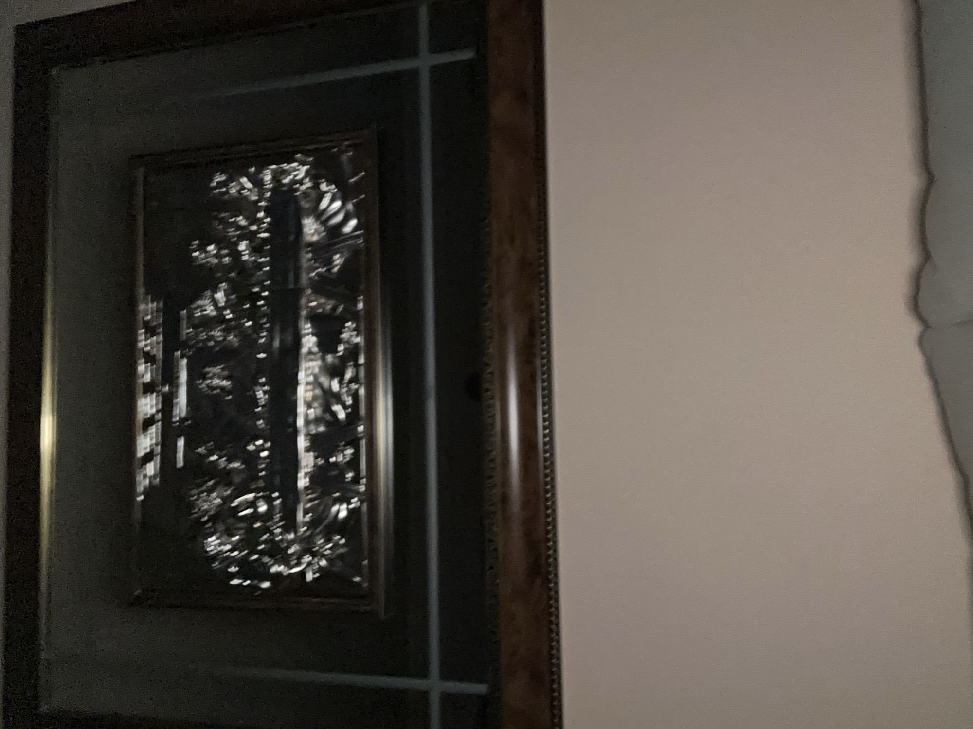 A framed art piece hangs on a wall with light pink paint. The frame is dark and ornate. The artwork within appears to be abstract and reflective. A light gray drape is partially visible on the right edge of the frame.