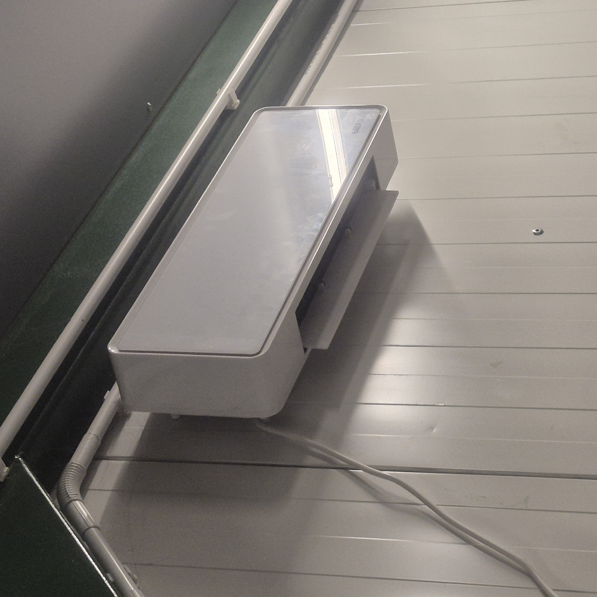 An indoor scene at an unknown location in Nowa Wieś Książęca, Poland, displays a wall-mounted air conditioning unit. The unit is white and rectangular, mounted high on a wall constructed of vertical, off-white panels. A white electrical cord extends downwards from the unit and is visible on the wall. Above the air conditioner, gray pipes are secured to a dark green band that runs along the top of the wall. The lighting in the scene is dim, casting shadows on the wall.