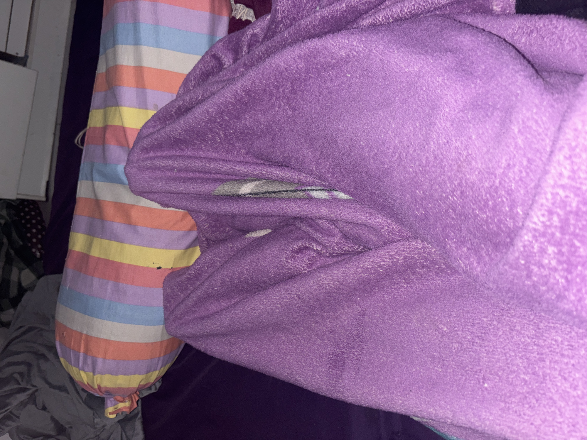In a dimly lit room, a close-up shot focuses on a person's legs covered by a plush, purple blanket. The blanket is draped in a way that suggests the person is lying down, possibly in a bed. To the left and behind the blanketed legs, a bolster pillow with colorful stripes of pink, yellow, light blue, and white is visible. The pillow appears slightly worn with a few dark specks on its surface. The background consists of rumpled gray bedding and a hint of a purple bedsheet. The overall impression is one of a private, indoor space, likely a bedroom. The specific location in North Jakarta, Indonesia, cannot be determined from the visual information provided.