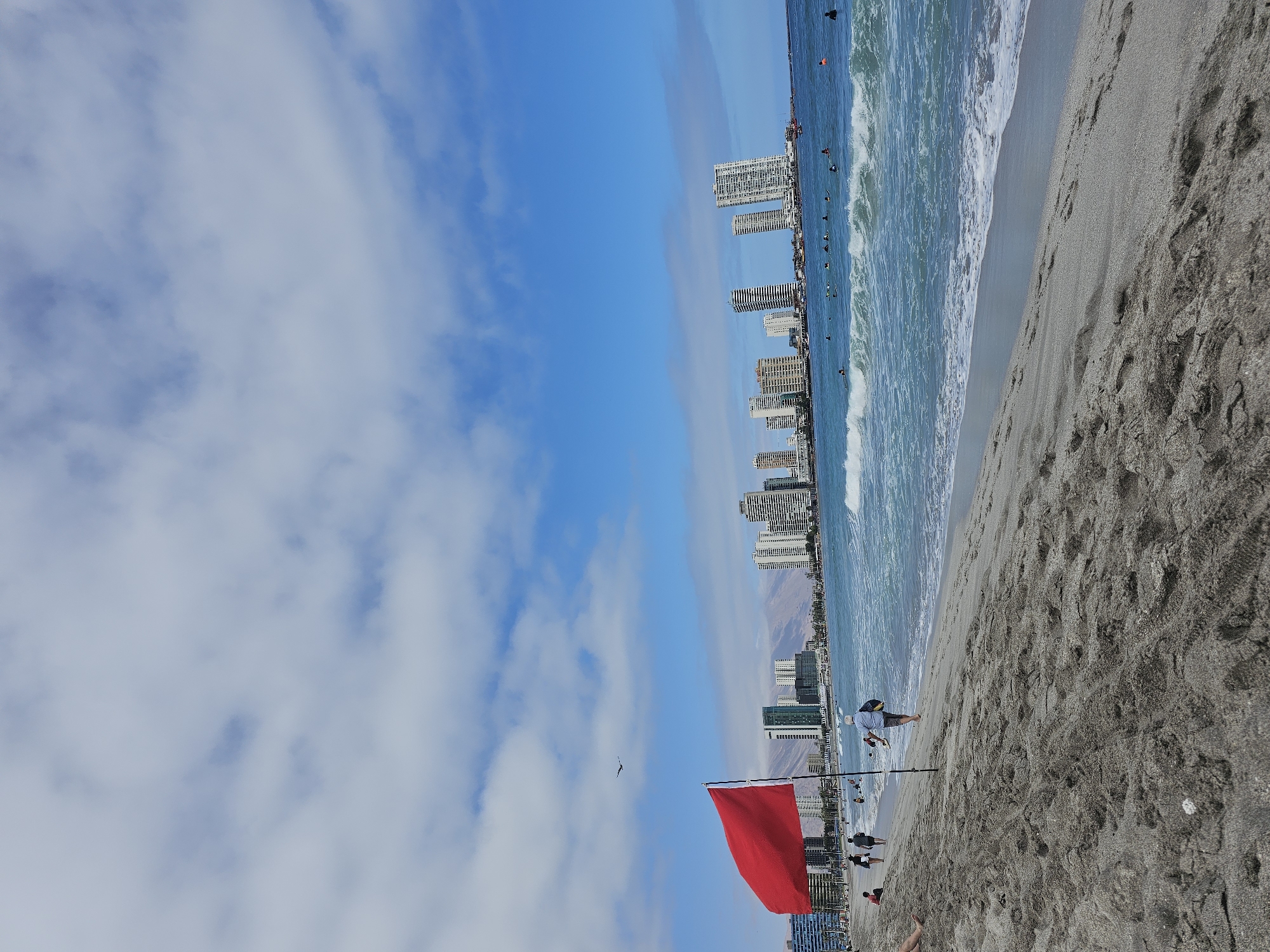 The image shows a beach scene in Iquique, Chile, with the cityscape in the background. A bright red flag, likely indicating a warning for swimmers, is prominently displayed on the left side of the foreground. The beach itself is sandy, with footprints visible. Several people are scattered across the sand and in the ocean. Some are walking along the shoreline, while others are swimming or surfing. The ocean is a vibrant turquoise color, with waves breaking near the shore. In the distance, a line of tall buildings forms the urban skyline of Iquique, with mountains visible behind the city. The sky is partly cloudy with a bright blue sky visible in between. A bird is seen flying in the sky above the red flag. The overall atmosphere appears to be a typical day at the beach with a clear indication of caution due to the red flag.