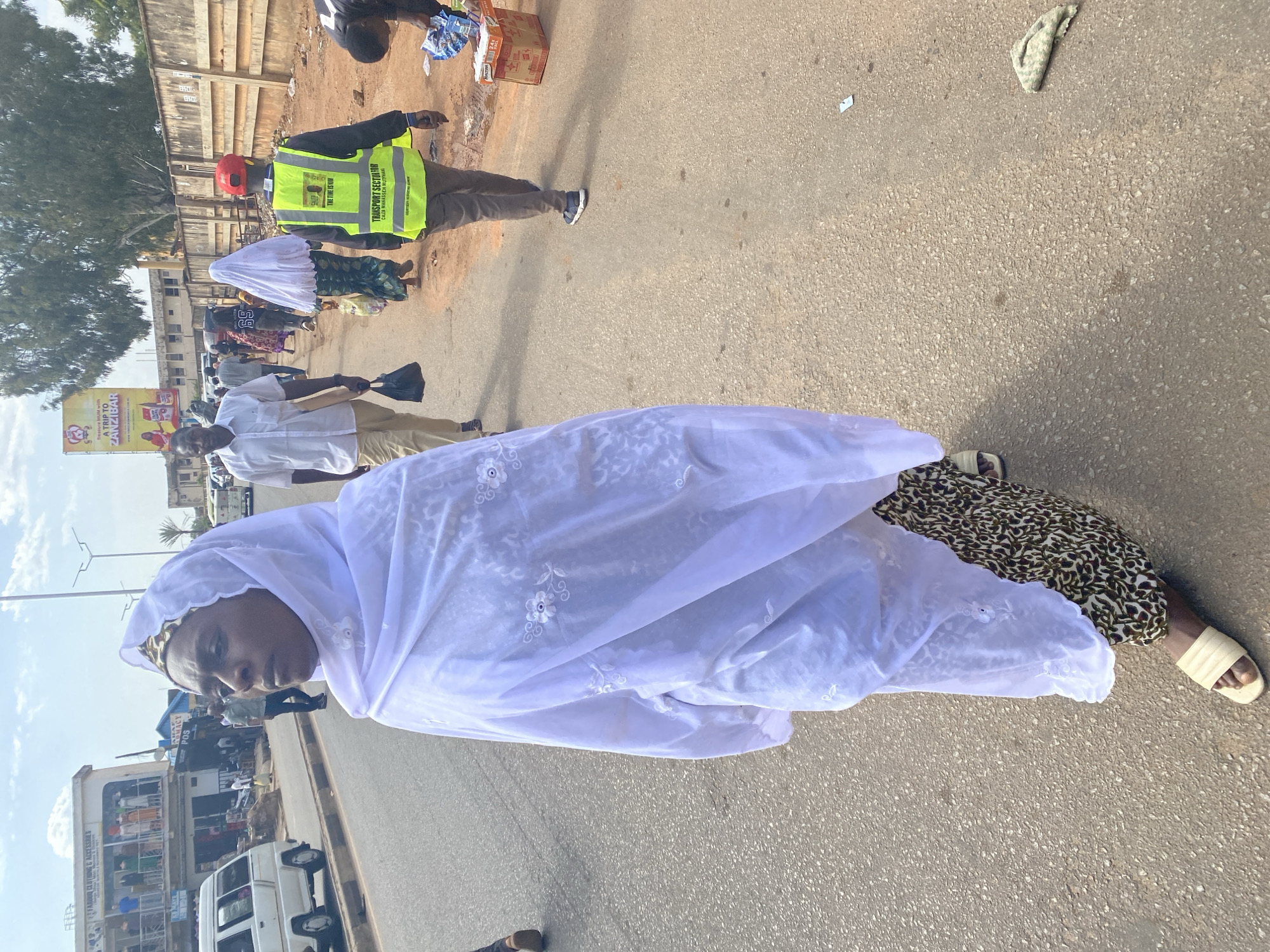 A woman in a white headscarf and patterned skirt is walking on a paved road in Jos, Nigeria. Behind her, a man in a fluorescent vest stands near other individuals. Shops and buildings line the background, including one with a sign for "FAROUD CLOTHING & ACCESSRES". A billboard advertising a trip to Zanzibar is visible. The road appears to be a main thoroughfare with traffic and pedestrian activity.