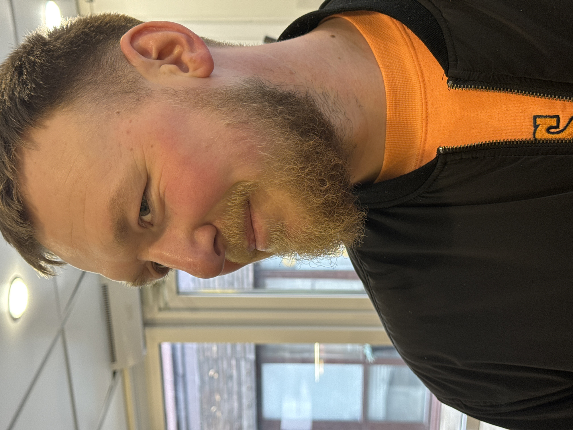 The image displays a close-up portrait of a man, originally rotated 90 degrees counter-clockwise. He has a reddish complexion, short, dark hair with a fade on the sides, and a light brown/blonde beard and mustache. He looks slightly downwards and to his right, with a subtle, relaxed smile.
He is dressed in an orange crew-neck t-shirt worn under a black zippered jacket, which is partially unzipped. A yellow design or logo, resembling part of a stylized letter "R," is visible on the orange shirt near the zipper.
The scene is indoors, with a large window in the background showcasing reflections and blurred outlines of an urban environment, suggesting buildings outside. A circular ceiling light fixture and a white wall with what appears to be a ventilation unit are also visible in the background. The lighting combines artificial light from the room and natural light filtering through the window. The overall atmosphere is casual, and the man appears comfortable. The location context is Perm, Russia.