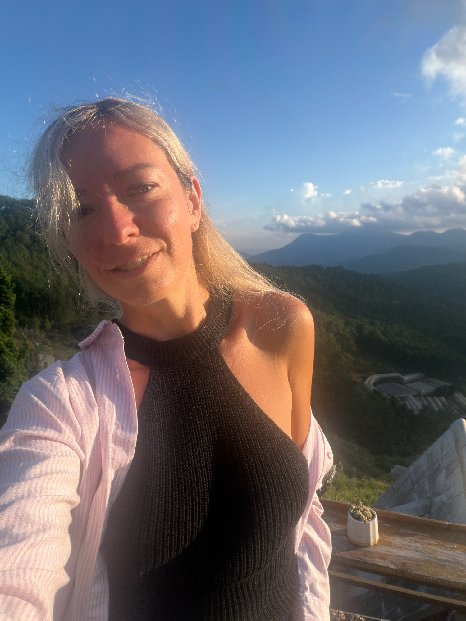 A blonde woman is taking a selfie outdoors with a mountainous landscape in the background, under a clear blue sky with some scattered clouds. She is wearing a black halter top and a light pink and white striped button-up shirt. The sun is shining, casting a warm glow on her face and the surrounding vegetation. The landscape appears to be a lush, green valley with several terraced fields or structures visible in the distance. The location is identified as Xuân Thành, Vietnam.