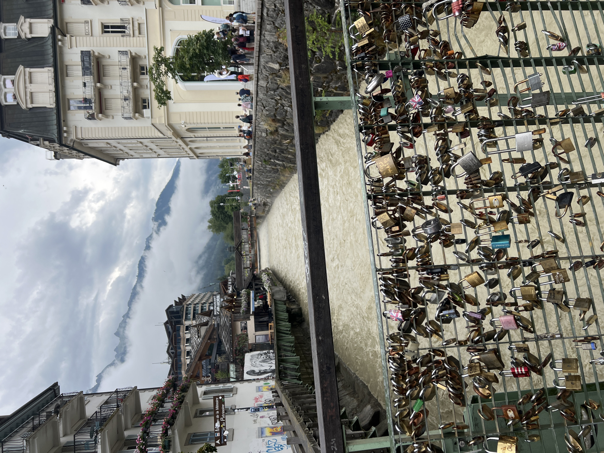The image depicts a wide river flowing through a town, with buildings lining the banks. In the foreground, a metal railing is covered with numerous padlocks, many of which appear to be love locks. The river water is a murky, light brown color and is moving swiftly. On the right side of the river, a large, ornate building with multiple stories and balconies dominates the scene. On the left side, there are more buildings, some with balconies decorated with flowers. In the background, mountains are visible, partially obscured by clouds. A group of people are seen gathered on a walkway along the riverbank, on the right side. The overall atmosphere is one of a bustling town situated in a mountainous region. The scene is likely Chamonix-Mont-Blanc, France, a popular tourist destination known for its alpine scenery and river.