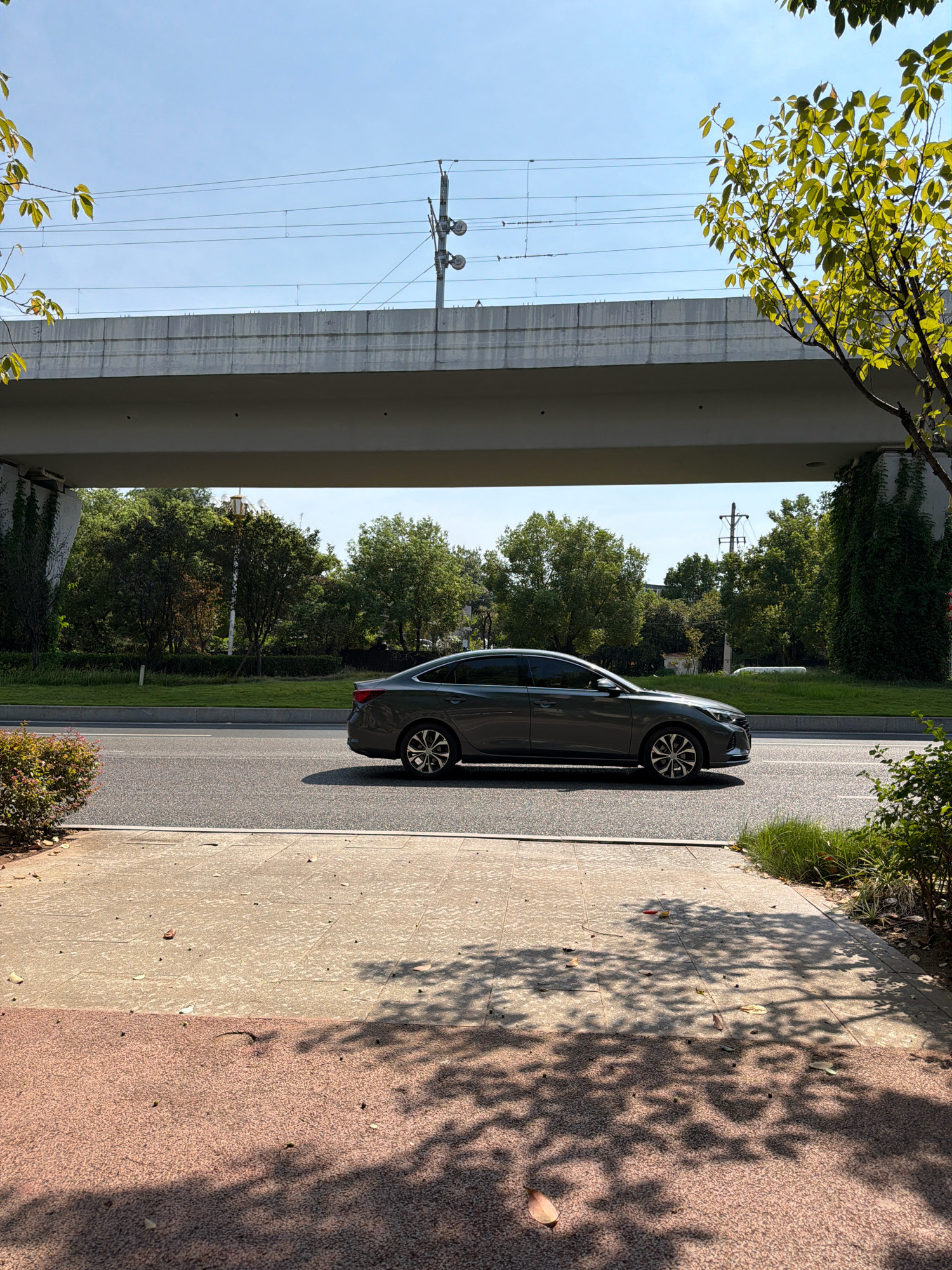 A gray sedan is driving on a road under an elevated structure. The elevated structure appears to be a bridge or overpass, and overhead power lines are visible. Trees and green spaces line the road and surround the elevated structure. The foreground shows a sidewalk with shadows cast by trees. The image is taken on a sunny day with a clear blue sky.
