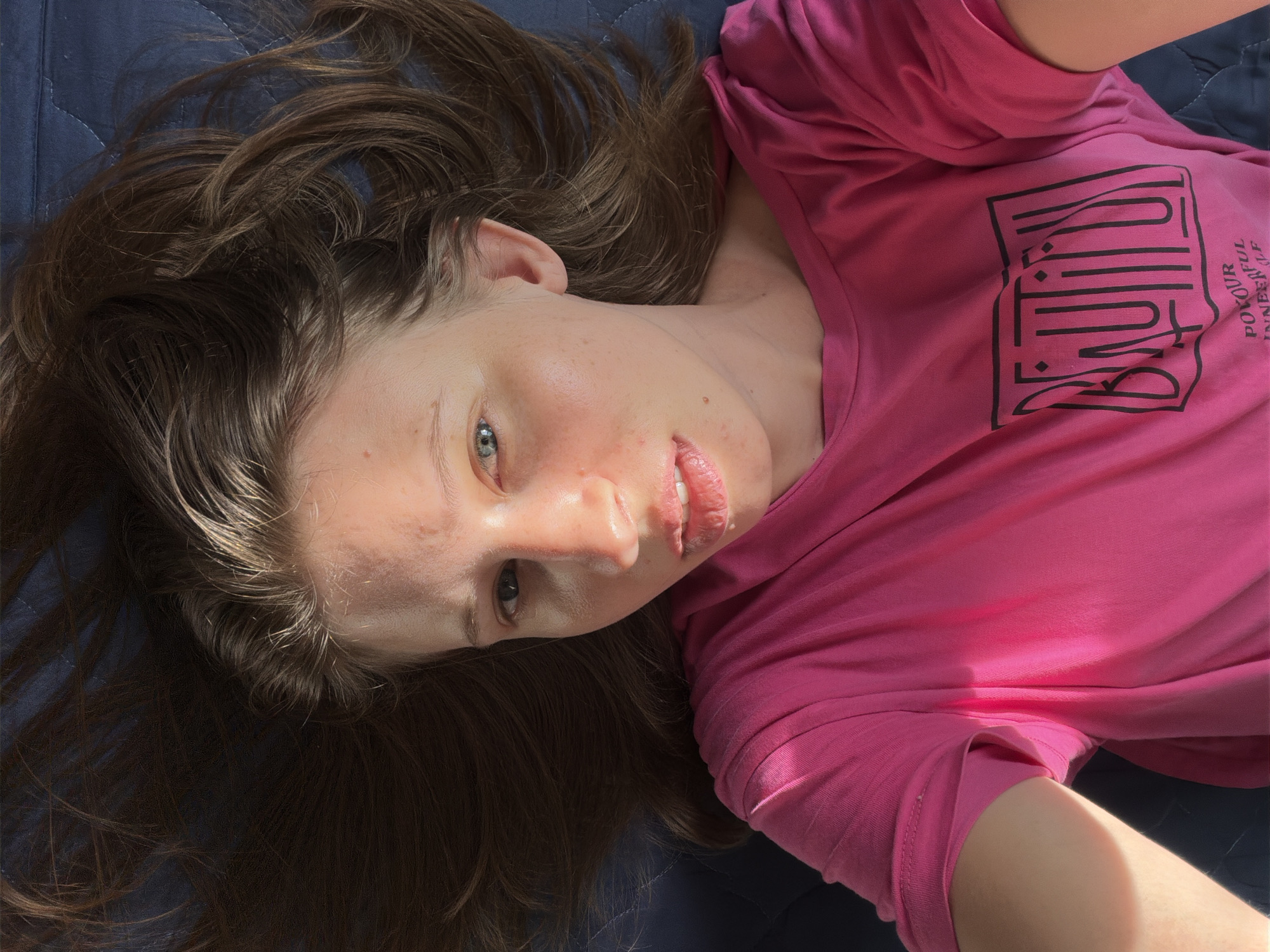 The image shows a close-up, overhead view of a young person lying on a dark blue surface, possibly a bed or sofa, with their head tilted to the right. The person has long, brown, wavy hair spread out around their head and shoulders. They are wearing a bright pink t-shirt with a graphic design in black. The design appears to be text within a rectangular frame, with additional text below it. Some of the visible text reads "BEAUTIFUL" and "POYOUR INNERFUL".
The person's face is partially visible, with their eyes looking towards the camera. Their lips are slightly parted, and they appear to have some minor skin imperfections, such as blemishes. The lighting suggests it might be daytime, with some bright sunlight casting shadows on the t-shirt. The overall impression is one of casual relaxation. The setting appears to be indoors, likely a bedroom or living area. No specific time of day or weather conditions can be definitively determined, beyond the presence of bright light.
