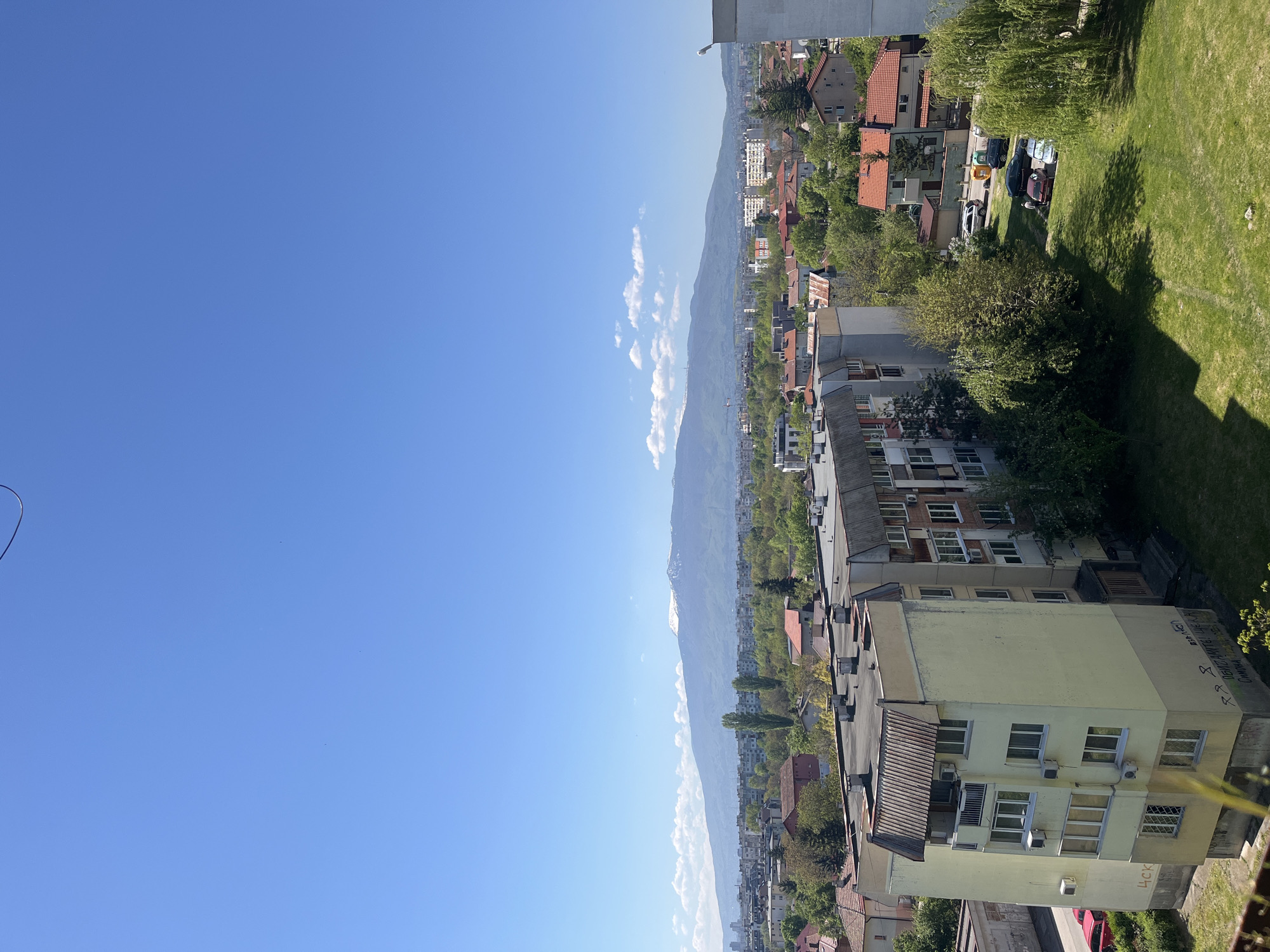 The image shows a view of Sofia, Bulgaria, looking from a high vantage point, likely an upper floor of a building. The right side of the image shows a mixed urban landscape consisting of residential buildings with a mix of old and newer construction. There are also signs of green spaces with trees and grassy areas interspersed among the buildings. A mountain range can be seen on the horizon. The sky occupies the left side of the image, revealing a clear, sunny day.