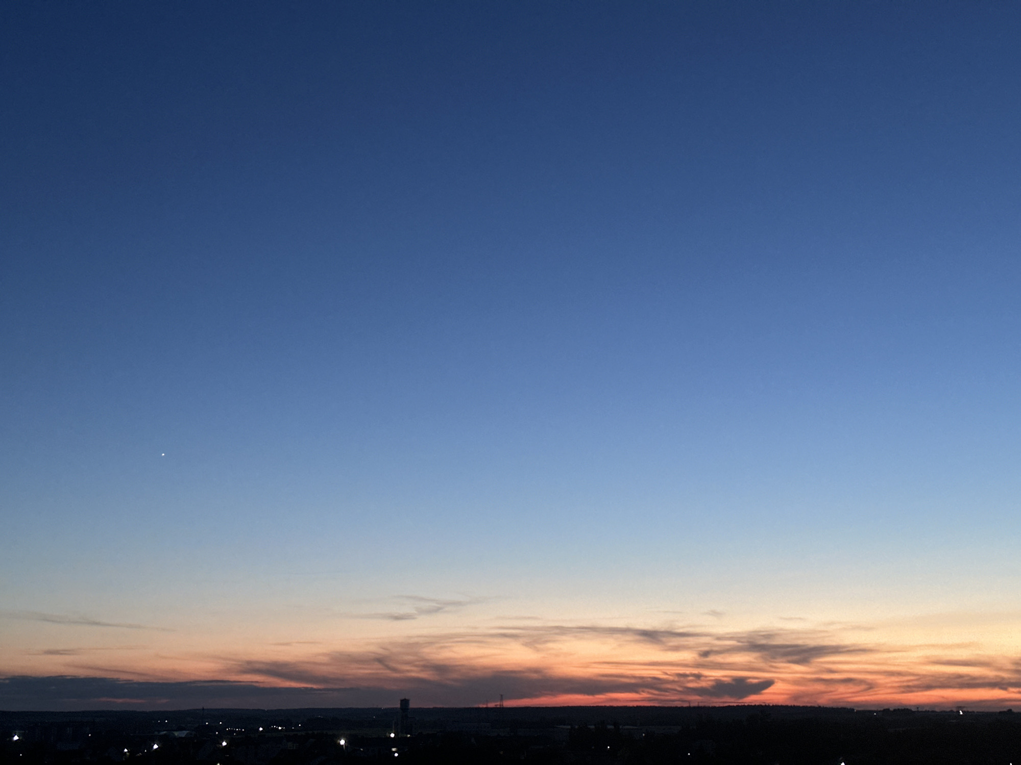 The image depicts a twilight sky over the city of Shchomyslitski Sel'ski Savet, Belarus. A clear gradient of blue dominates the upper portion of the sky, transitioning to lighter hues of blue and pink near the horizon. Wisps of orange and red clouds are visible at the lowest part of the sky, indicating sunset. A single, bright star is visible in the upper left quadrant of the sky. The foreground is dark, with the silhouettes of buildings and scattered lights suggesting an urban or semi-urban environment. The overall scene captures the serene beauty of the sky at dusk.