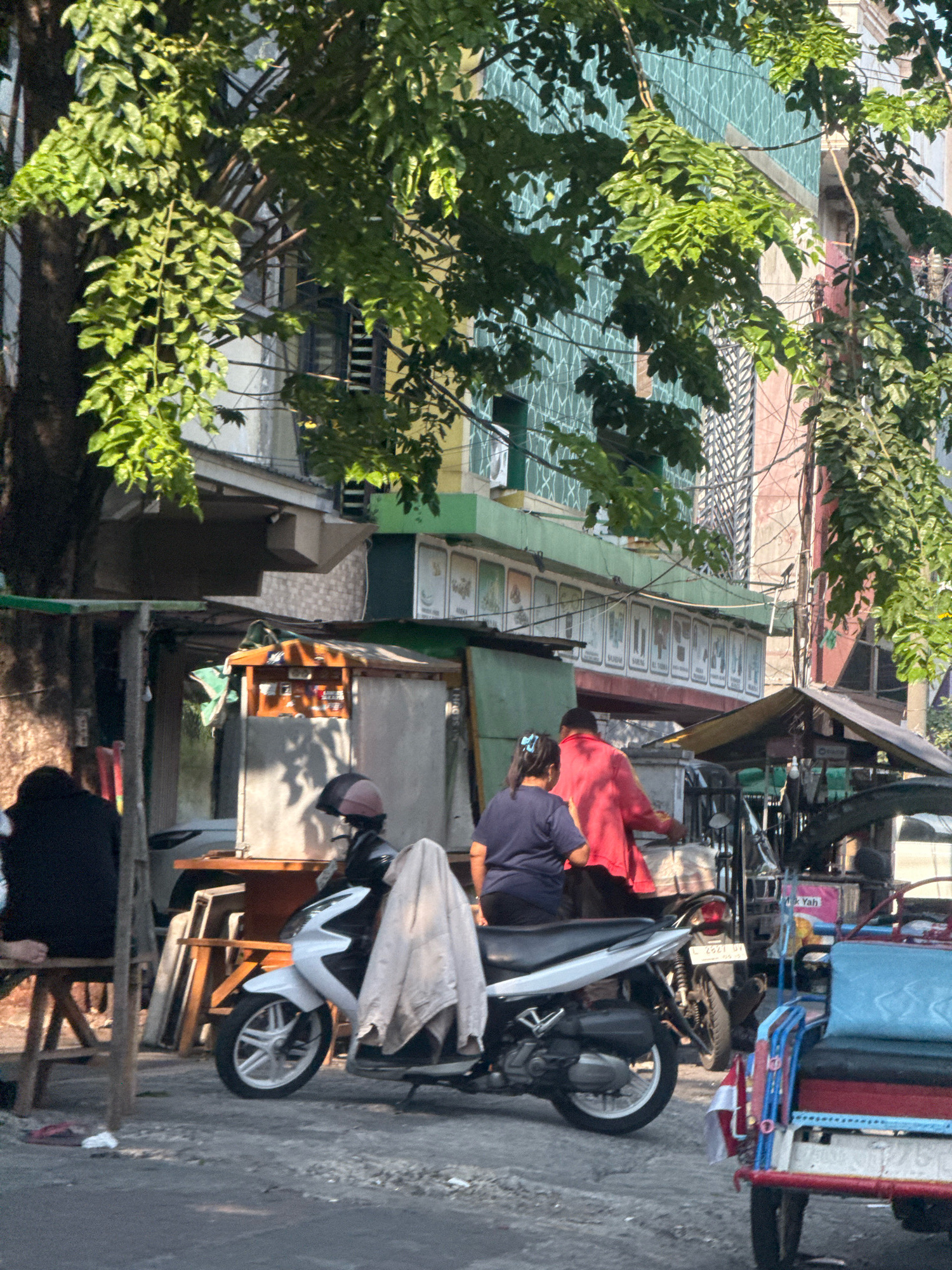 The image captures a street scene in Surabaya, Indonesia, on a sunny day. In the foreground, a white scooter is parked, draped with a light-colored garment. To the left, a person in dark clothing sits on a wooden stool. Further back, two individuals are walking; one in a red shirt and another in a dark blue shirt with a ponytail. Behind them, a street vendor's cart with a brown wooden structure and white panels is visible. The background features buildings with green and blue tiled facades, some with arched windows and electrical wires strung across. A small red and blue vehicle, possibly a tricycle or a pedicab, is parked on the right side of the frame. The ground is paved with gravel and asphalt. Lush green foliage from trees overhangs the scene, casting shadows.