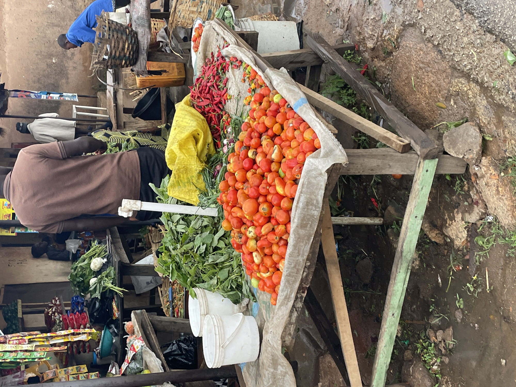 The image depicts a market scene in Jos, Nigeria. A large pile of red tomatoes dominates the foreground of a makeshift wooden stall covered in white fabric. To the left of the tomatoes are red chili peppers, a yellow cloth, green onions, leafy greens, and two white plastic buckets. A man in a brown shirt stands partially visible behind the stall. In the background are additional stalls with merchandise, and people are moving about. The ground is uneven and muddy.