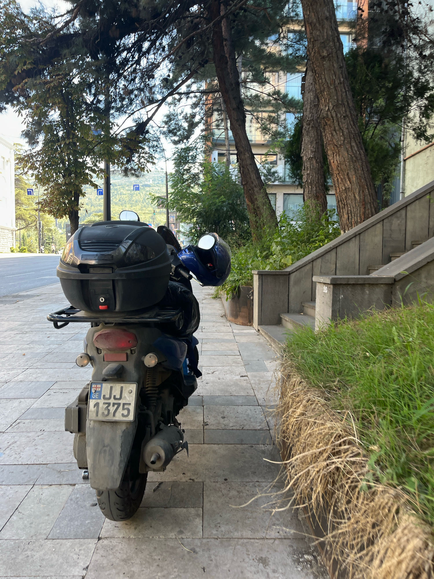 A dark blue scooter is parked on a paved sidewalk. The scooter has a black top case, a blue helmet attached to the handlebars, and a license plate that reads "GE JJ 1375". In the background, there is a modern building with multiple floors and windows, surrounded by trees and greenery. A concrete staircase with railings is visible to the right of the scooter, leading upwards. The sidewalk is made of large, grey rectangular tiles. The overall setting appears to be an urban environment in Tbilisi, Georgia, likely on a street with some elevation changes given the presence of stairs and a view of distant hills.