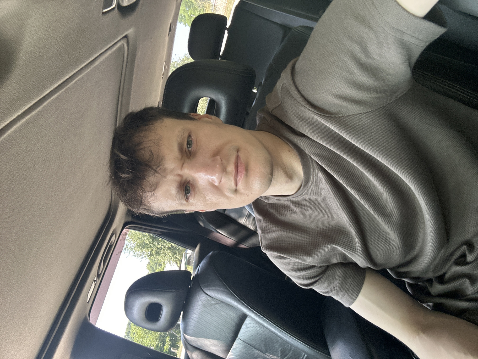 A man is sitting in the driver's seat of a car. He is looking at the camera and has a neutral expression on his face. The car's interior is visible, with black leather seats and a sunroof. Outside the car, trees and a slightly overcast sky can be seen. The image is taken from a slightly elevated angle.