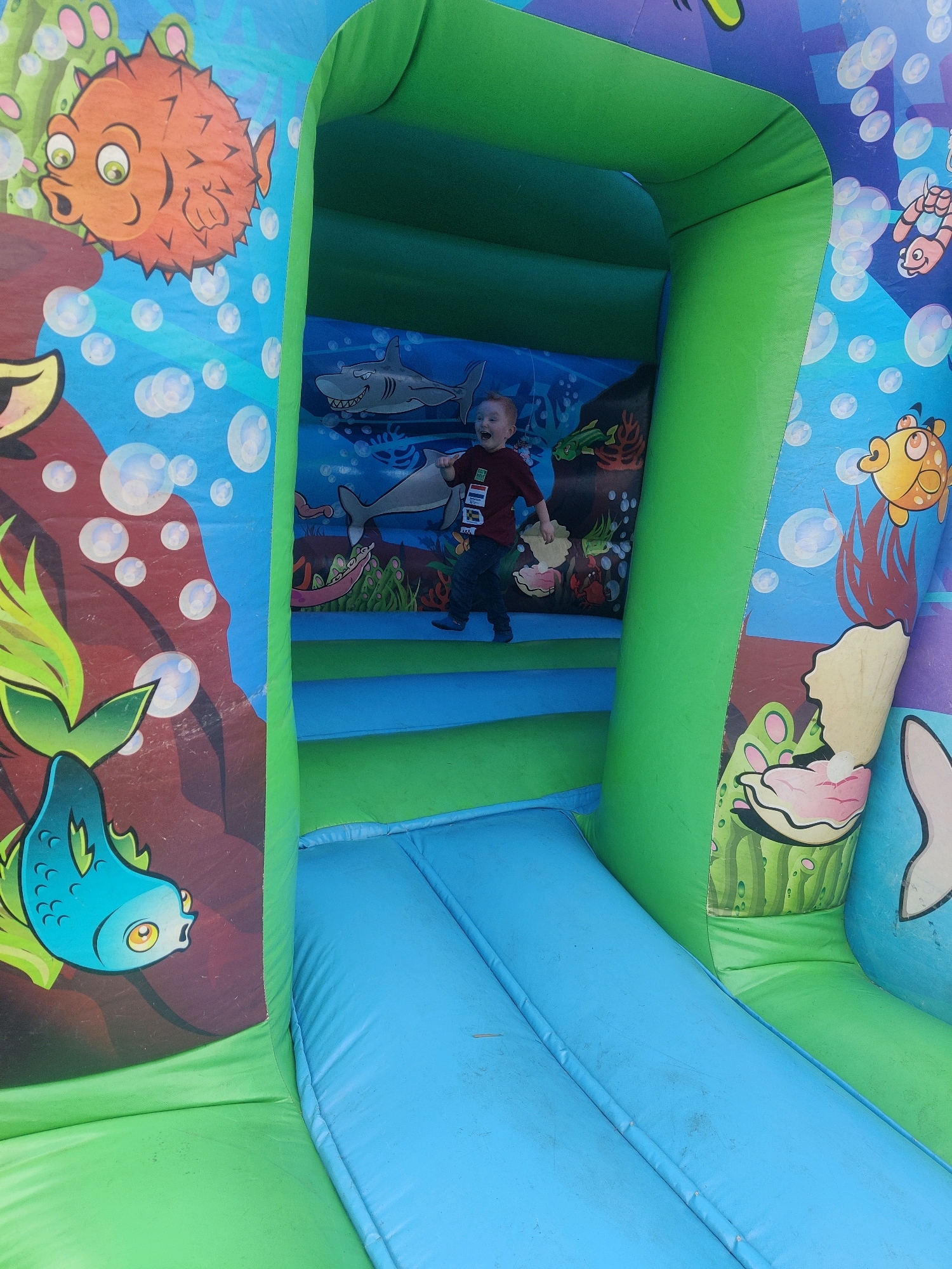 In Tilburg, Netherlands, a young boy with red hair is captured mid-jump inside a vibrant, inflatable castle adorned with an underwater theme. He is wearing a dark red shirt with various patches and dark pants. The castle walls are decorated with cartoonish sea creatures, including a pufferfish, sharks, and a blue fish. Bubbles are depicted throughout the background, contributing to the aquatic setting. The boy is positioned on an elevated blue platform, with a bright blue slide extending downwards from his right. The inflatable structure is primarily green and blue, with other colors appearing in the artwork. The image has a slightly gritty texture, suggesting it was taken outdoors.