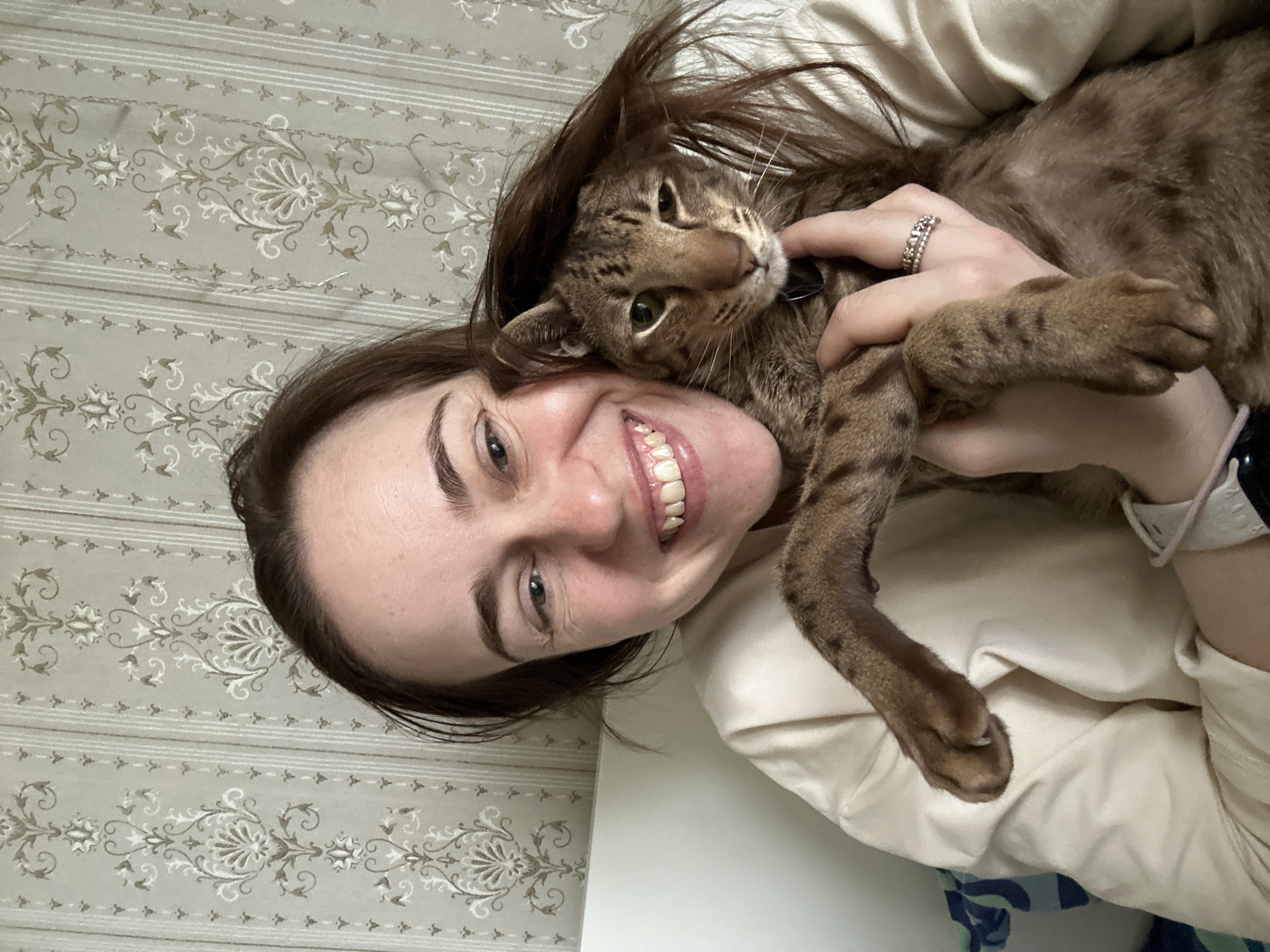A young woman is captured in a close-up, horizontal portrait, smiling broadly at the camera while embracing a cat. She has brown hair and is wearing a light-colored, possibly cream, top. A silver-toned ring is visible on her left hand, and a light-colored watch is partially seen on her right wrist.
The woman is lying down, and a brown and gold tabby cat is snuggled against her face and chest, looking directly towards the viewer with green eyes. Her arm is around the cat, holding it affectionately. The cat appears calm and content.
The background consists of a patterned surface, resembling wallpaper or bedding, featuring light grey horizontal stripes alternating with ornate floral and vine motifs in lighter shades. The scene conveys a warm and joyful moment of connection between the woman and her pet. The exact time of day is not indicated, and while the context of Moscow, Russia, is provided, there are no specific visual cues in the image to confirm this location. No visible text is present.