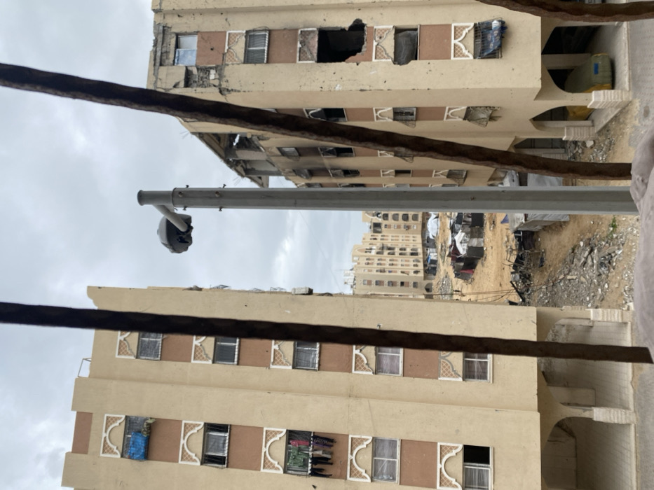 This image depicts an urban landscape in Khan Yunis, Palestinian Territory, framed by two vertical, dark brown, out-of-focus bars in the foreground. An overcast sky dominates the upper portion.
In the midground, a gray utility pole with a mushroom-shaped fixture (possibly a camera or lamp) stands centrally. To the left, a multi-story, light beige apartment building with brown vertical strips and white arched window accents shows signs of damage, specifically a missing section from its top-right facade. Laundry hangs from a lower-level window, and a blue tarp covers an opening on an upper floor. The ground floor features arched open passages.
To the right, another similar multi-story building exhibits extensive damage, including large holes in its facade, exposed rebar, and burn marks around several window openings on its upper floors. Windows are broken or missing. A yellow object, potentially a covered vehicle, is partially visible within a ground-floor arched passage.
Between these two foreground buildings and several similar, less-damaged structures in the background, lies a large, open, sandy area. This area contains scattered piles of rubble and debris. A cluster of makeshift shelters, constructed from tarps, corrugated metal, and other salvaged materials, forms a temporary encampment within this open space. Several small, indistinct human figures are visible near these shelters.