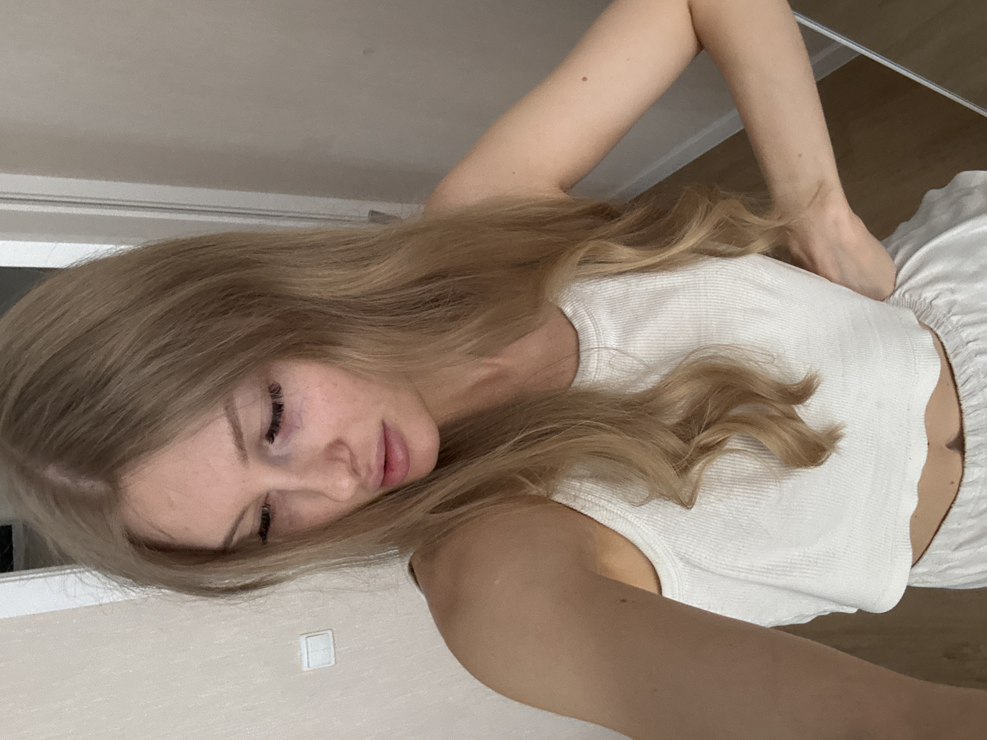 The image depicts a woman with long, blonde hair. She is wearing a white tank top and white shorts. She is lying down, and her right arm is raised behind her head, touching her waist. The background shows a beige wall with a light switch and a wooden floor. There is no information in the image that indicates the location is Perm, Russia.