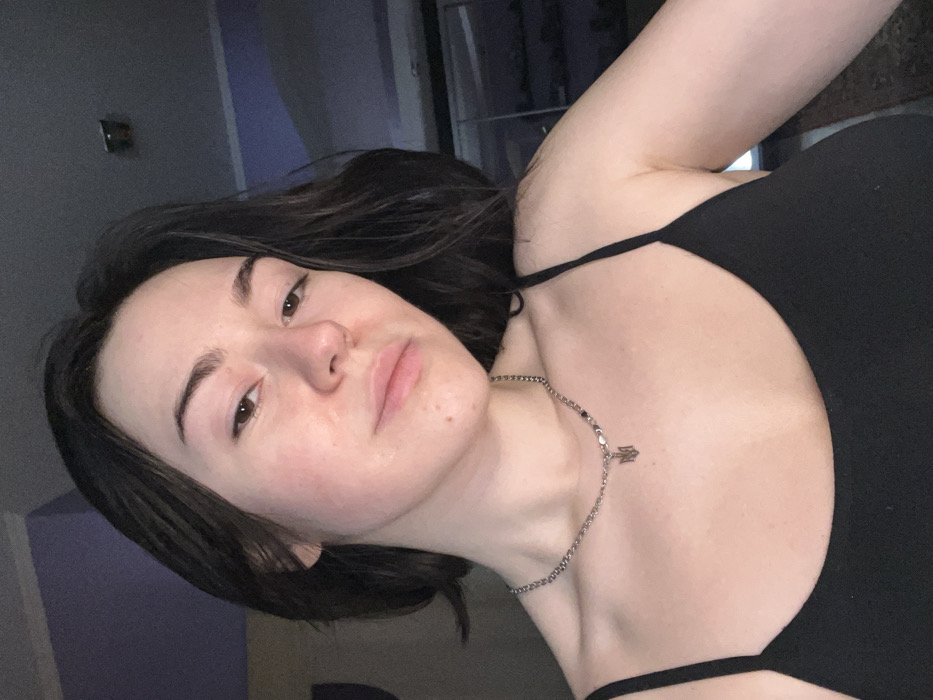 A young adult female, identified as residing in Odesa, Ukraine, is captured in a close-up, chest-up indoor portrait. She possesses dark, shoulder-length hair and fair skin, with a neutral expression as she gazes directly into the camera. She wears a black, spaghetti-strap camisole and a silver-toned chain necklace adorned with a pendant depicting the Ukrainian Tryzub. The background reveals an interior room with a purple-hued wall on the left and a darker, reflective surface on the right, suggestive of a mirrored furniture piece or window. Illumination is low and ambient, indicative of an indoor setting.