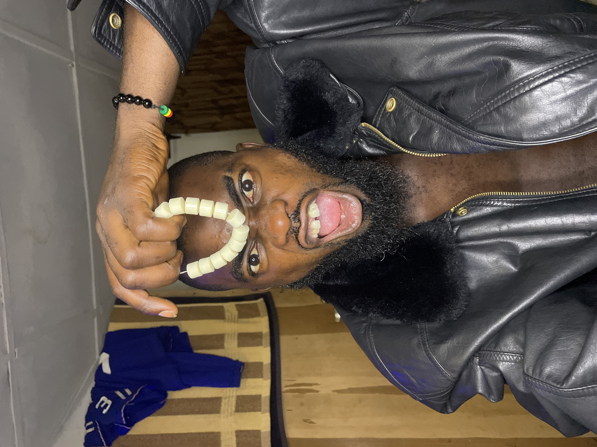A close-up, chest-up portrait depicts an adult Black male indoors. He has a dark complexion, a full black beard, and an intense expression with his mouth wide open, revealing his tongue and several teeth, including a visible gap between his upper front teeth. His left eye (viewer's right) is wide open. His right hand (viewer's left) is raised, holding a string of cream-colored, rectangular beads in an inverted U-shape above his forehead. He is wearing an open black leather-like jacket with a prominent black faux fur collar, exposing his bare chest. On his left wrist (viewer's right), a black beaded bracelet with red, yellow, and green accent beads is visible. The background on the left features a blue garment (possibly a jersey with a white number "3") hanging over a striped tan and brown fabric or wall covering. A white paneled ceiling is visible at the top. The specific location in Jos, Nigeria, cannot be determined from the visual information in the image.