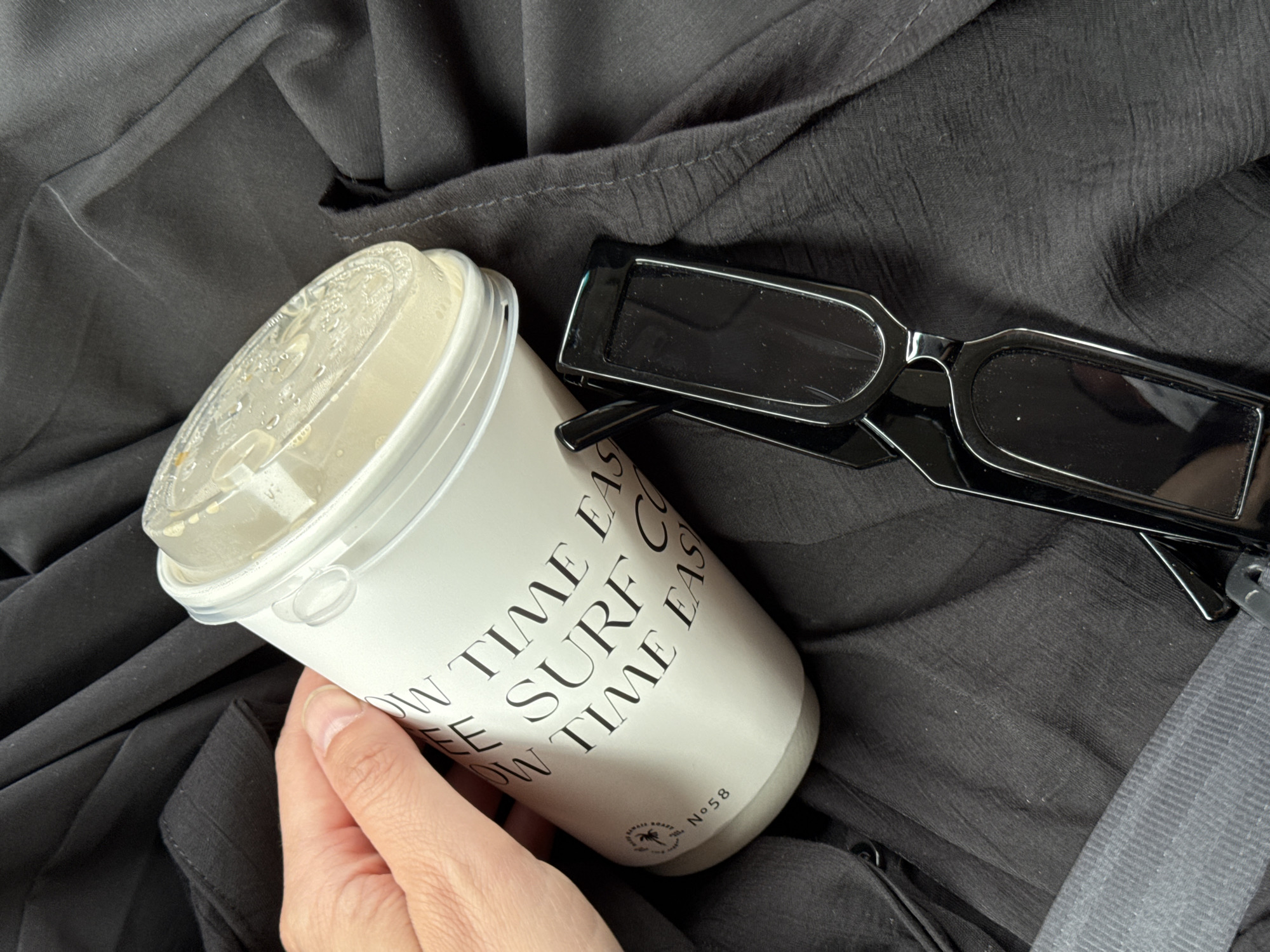 A human hand is holding a white paper coffee cup with a plastic lid. The cup has black text printed on it. Next to the cup sits a pair of black sunglasses on top of black fabric. A car seatbelt is visible in the lower right corner of the image. The location in the city of Alan-Polyan, Russia is unknown.